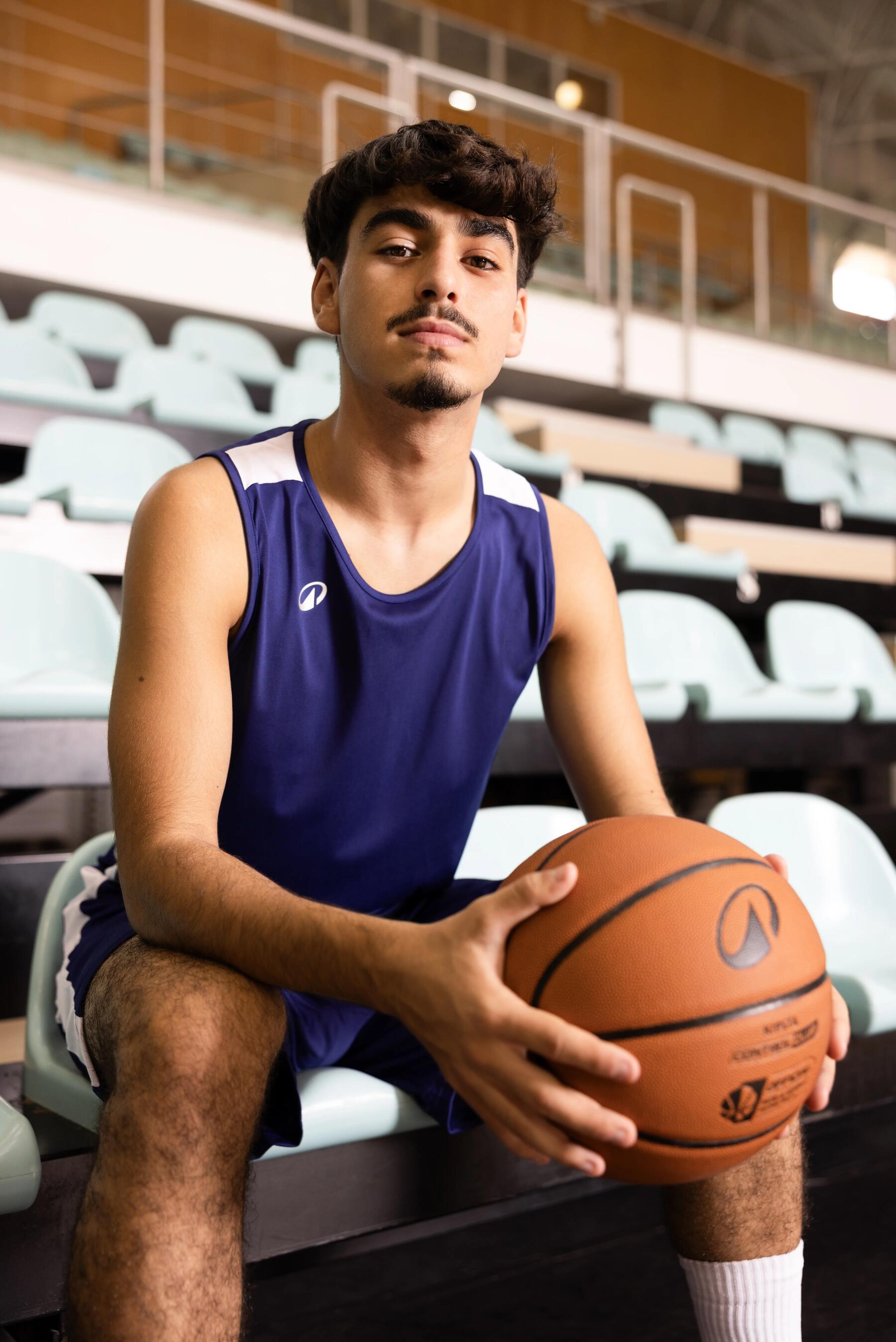 Ein junger Mann im blauen Basketballtrikot sitzt mit einem Basketball in den Händen auf den Zuschauerrängen einer Sporthalle.