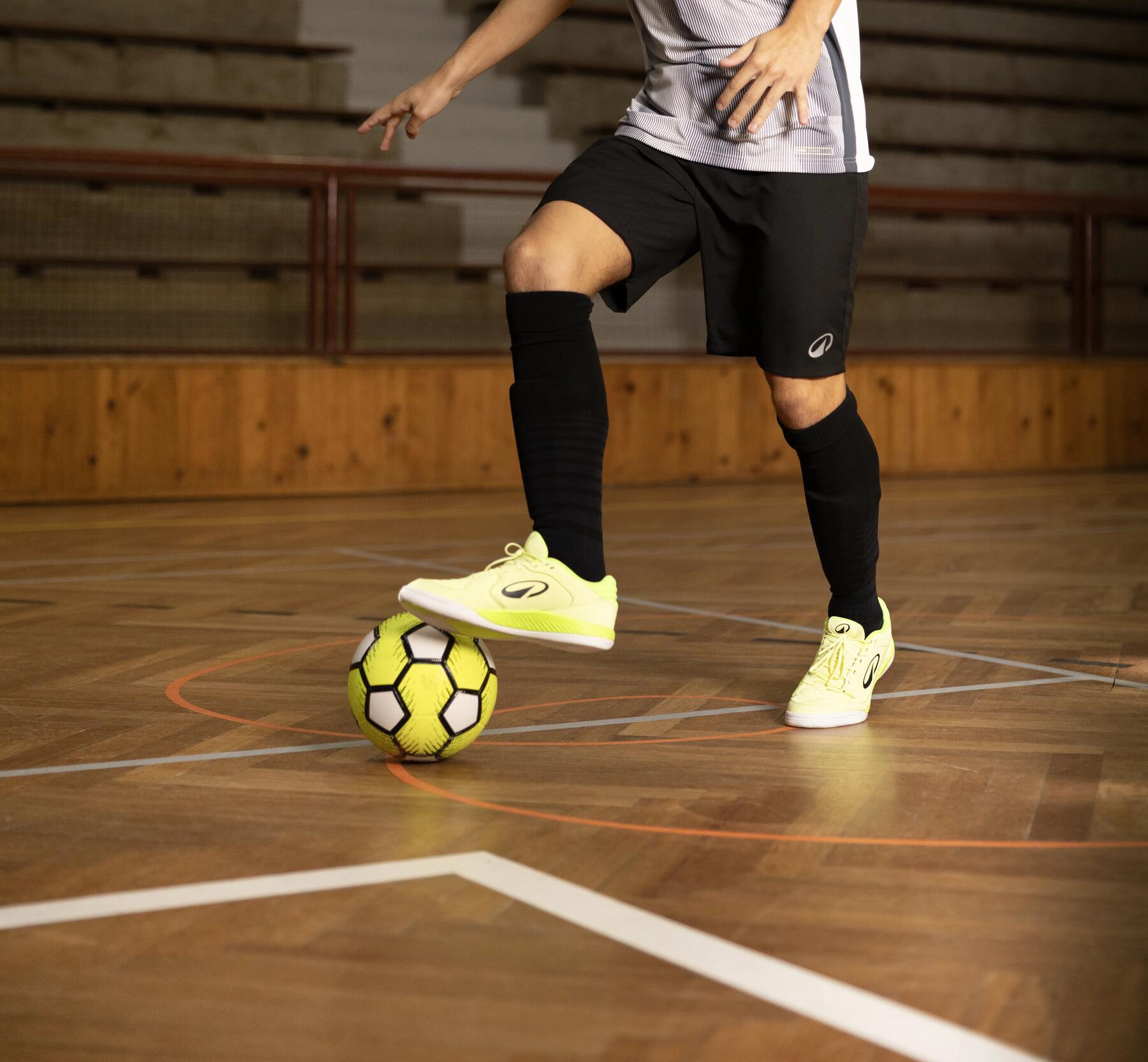 Ein junger Mann in grau-schwarzer Sportbekleidung kontrolliert mit leuchtend gelben Schuhen einen gelben Fußball auf dem Parkettboden einer Sporthalle.