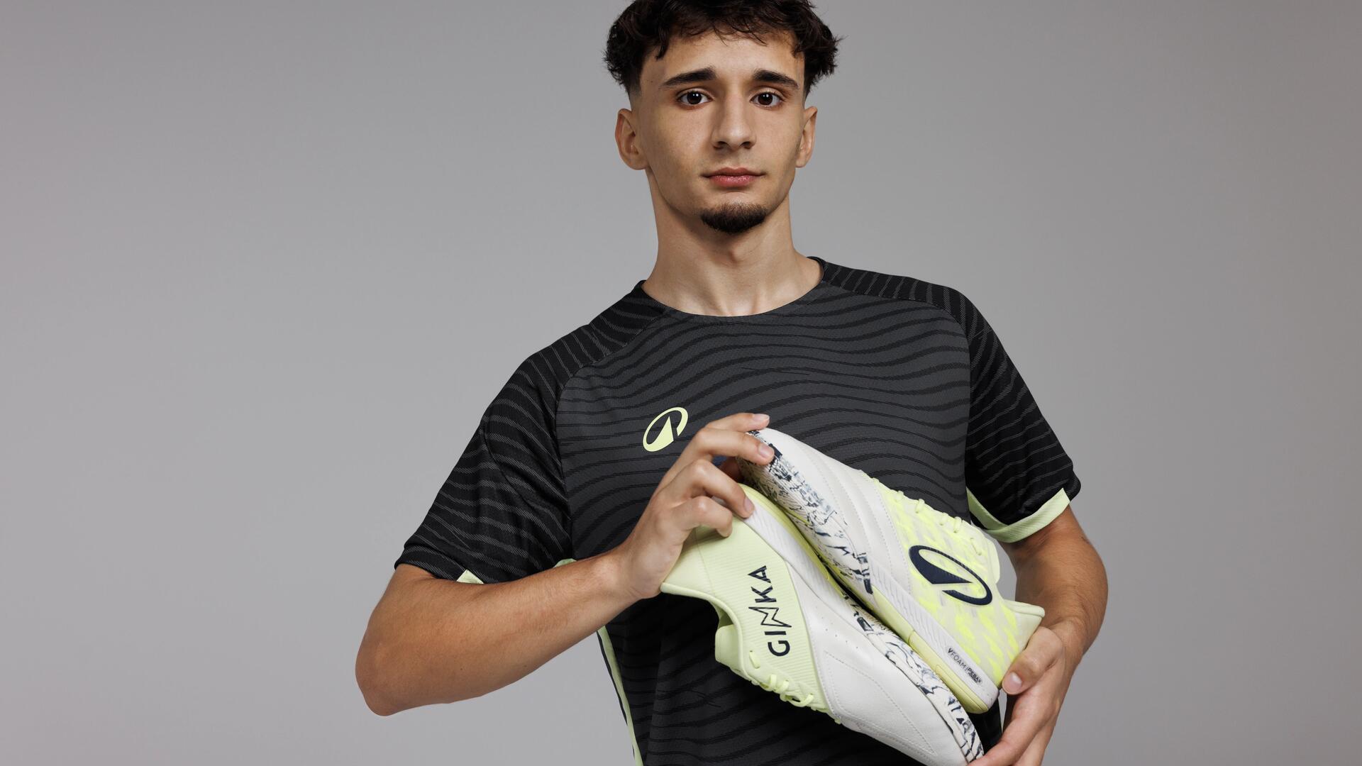 Atleta de futsal segura sapatilhas Ginka amarelas e brancas com padrão marmoreado sobre t-shirt técnica preta e cinzenta.