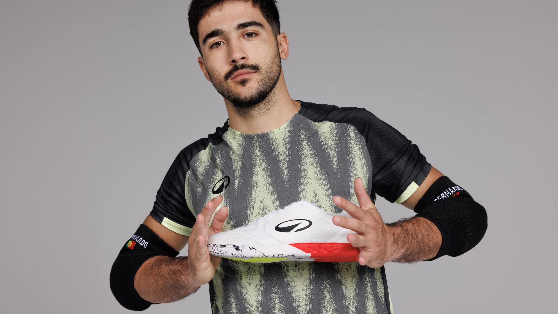 Jogador de futsal segura sapatilha desportiva branca com detalhes vermelhos da marca Kipsta, vestindo equipamento técnico cinzento e preto.