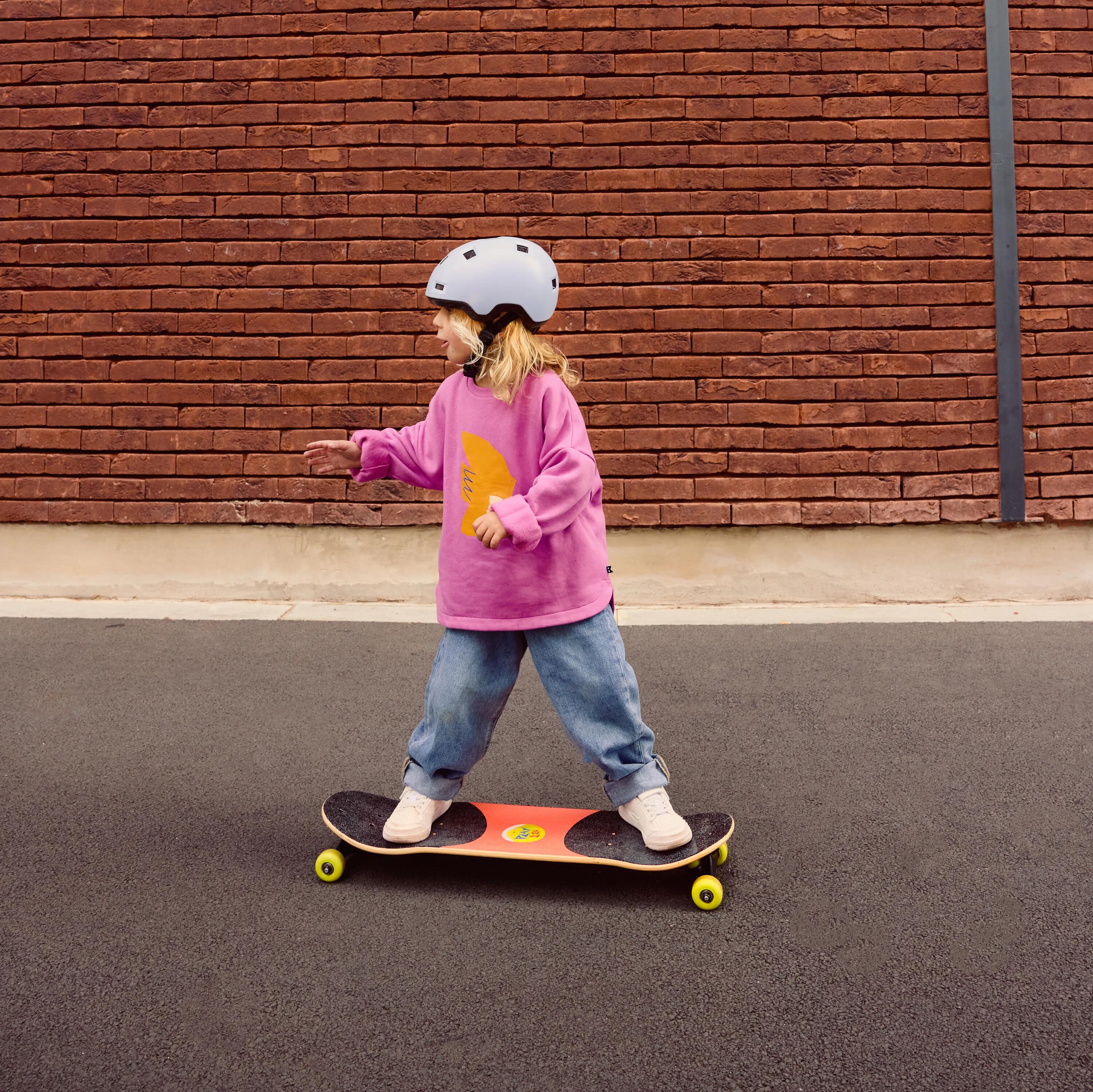 Seitenansicht eines Kindes mit hellem Helm und pinkem Pullover, das auf einem Longboard steht. Das Board hat schwarze Grip-Pads und gelbe Rollen. Das Kind hält die Balance mit ausgestreckten Armen vor einer roten Backsteinwand.
