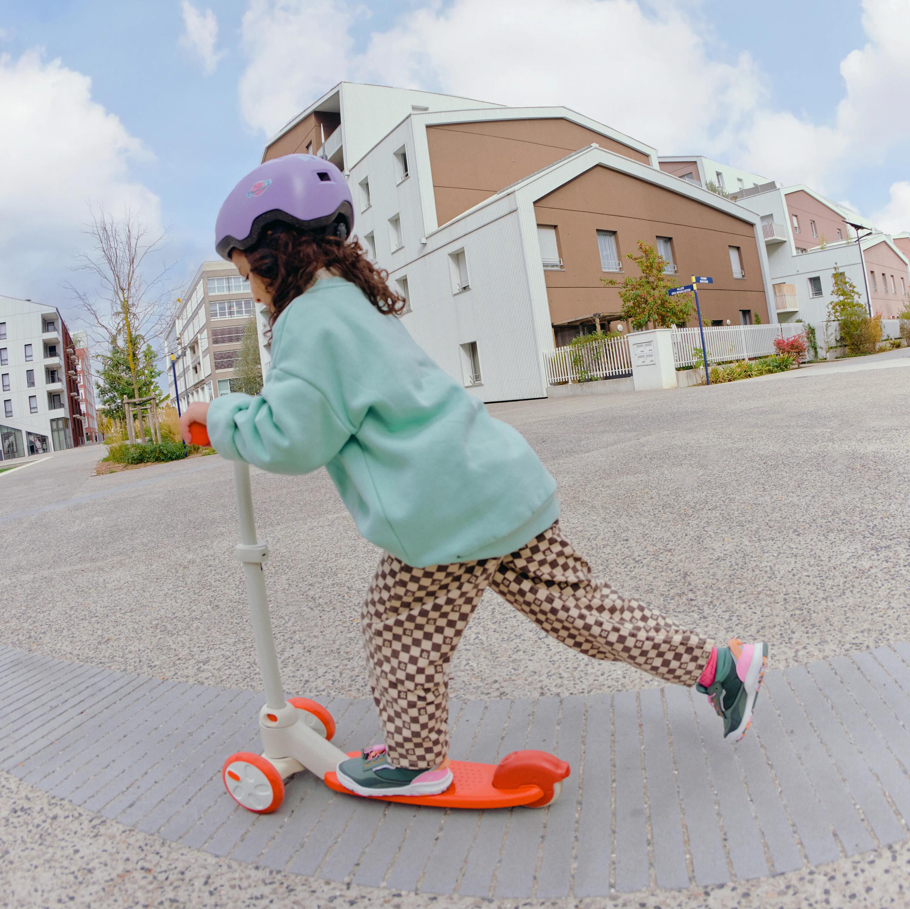 Ein Kind mit violettem Helm fährt auf einem hellbeigen Dreirad-Roller mit orangefarbenem Trittbrett über einen Platz.