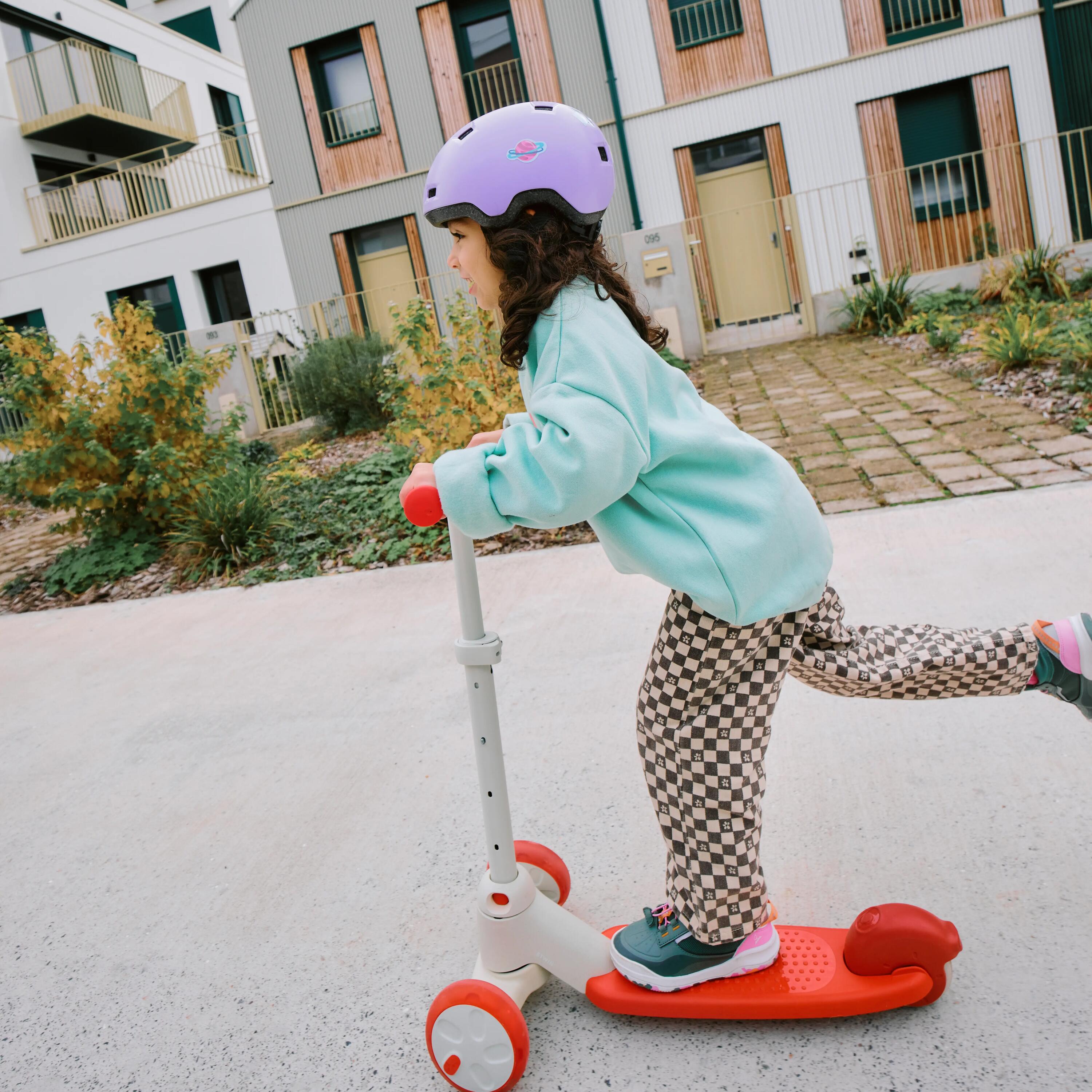 Junges Mädchen mit bunter Kleidung und lilanem Helm fährt einen beige-roten Roller auf einer Straße.
