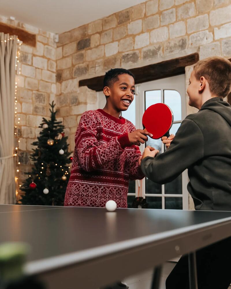 Zwei Jungen spielen lachend Tischtennis in einem weihnachtlich dekorierten Raum, wobei einer der beiden gerade seinen roten Schläger für einen Schlag bereit hält.