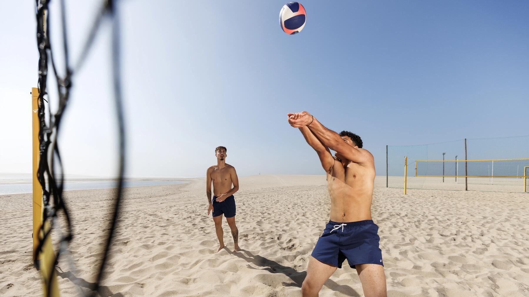 Zwei Männer mit oberkörperfreier Badehose spielen Beachvolleyball an einem sonnigen Sandstrand, während einer von ihnen den Ball pritscht.