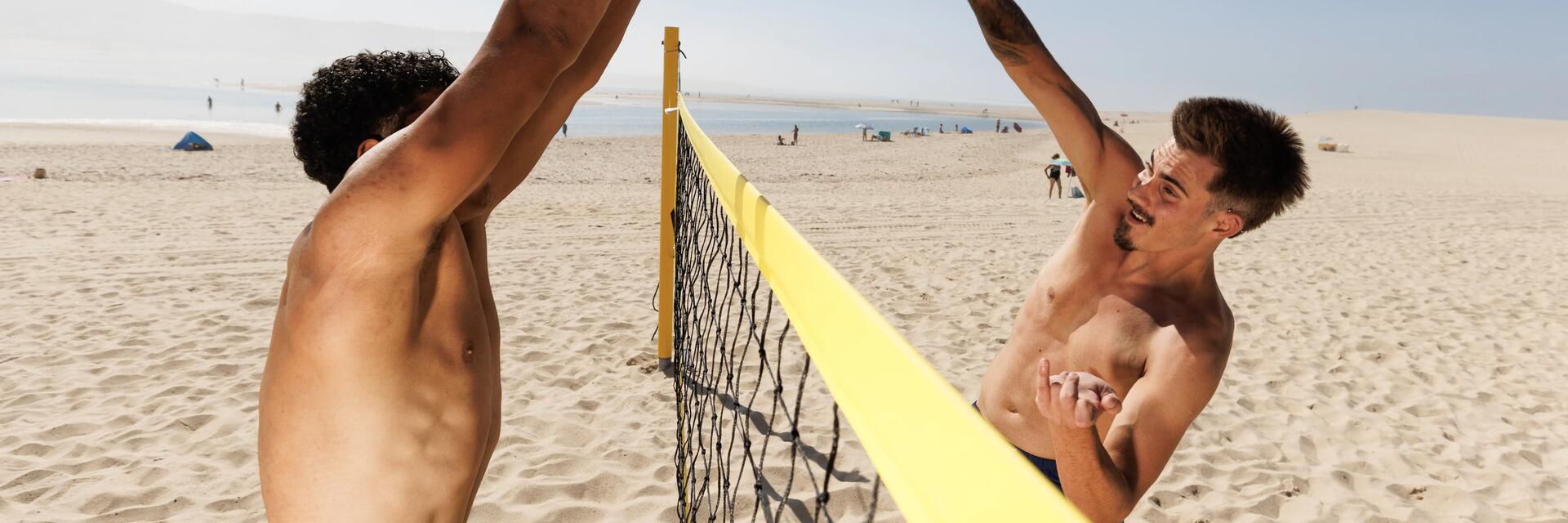 Zwei Männer mit freiem Oberkörper spielen an einem sonnigen Sandstrand Beachvolleyball über ein gelbes Netz hinweg.