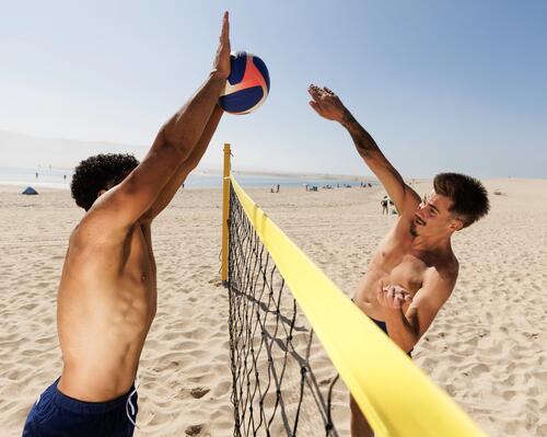 Zwei Männer mit freiem Oberkörper spielen an einem sonnigen Sandstrand Beachvolleyball über ein gelbes Netz hinweg.