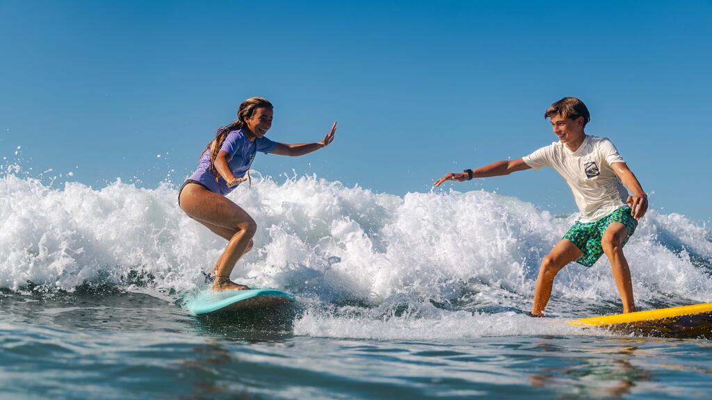 Alt-Text: Zwei Jugendliche surfen nebeneinander auf spritzenden Wellen im Meer. Links steht ein Mädchen in lila Shirt auf einem hellblauen Surfbrett, rechts ein Junge in weißem Shirt und grün gemusterten Badeshorts auf einem gelben Board. Beide halten lachend die Arme zur Ausbalancierung ausgestreckt, während im Hintergrund ein strahlend blauer Himmel zu sehen ist.