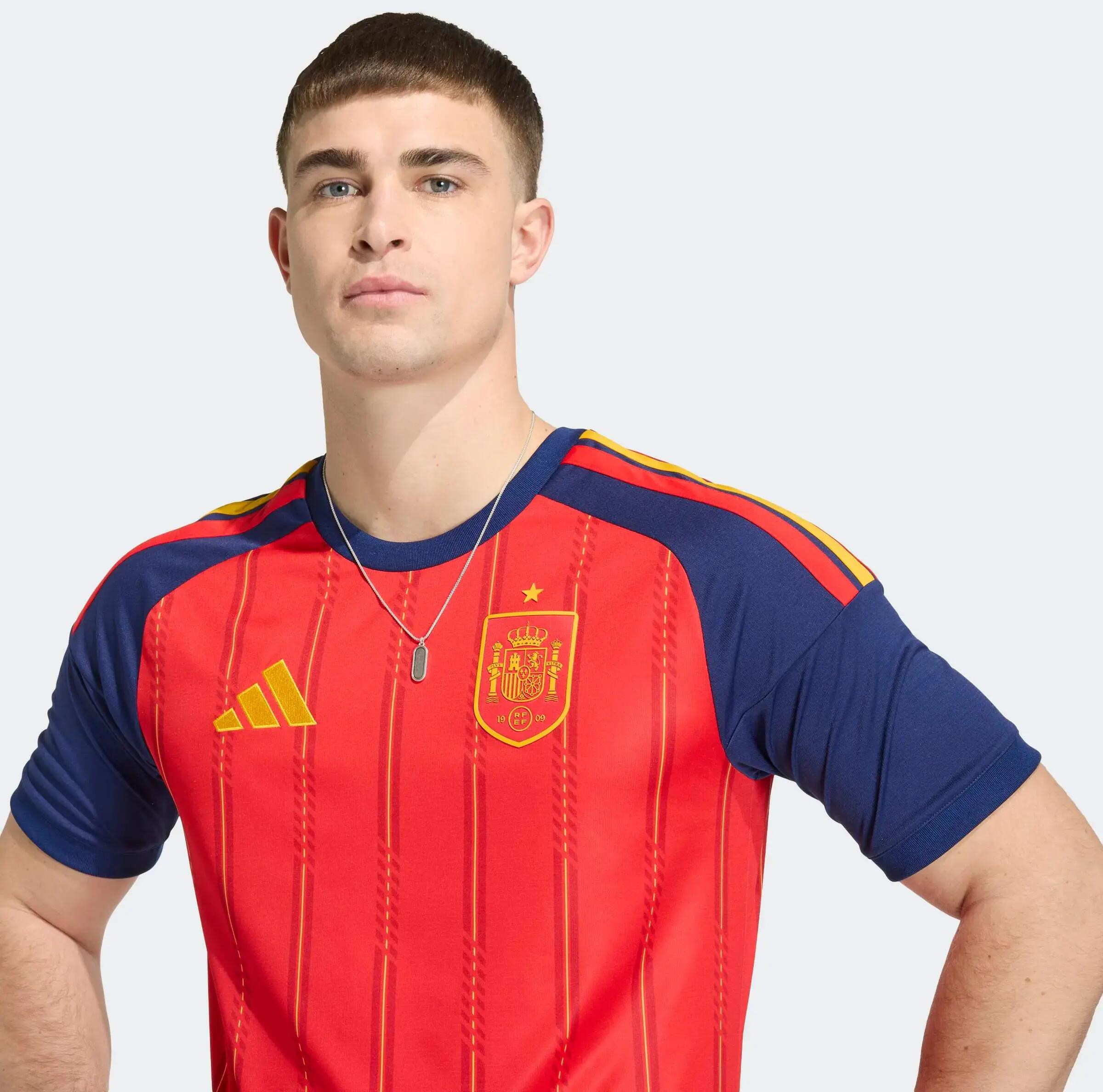 Ein Mann präsentiert das rot-blaue Spanien-Heimtrikot von Adidas mit gelben Akzenten und vertikalen Nadelstreifen.