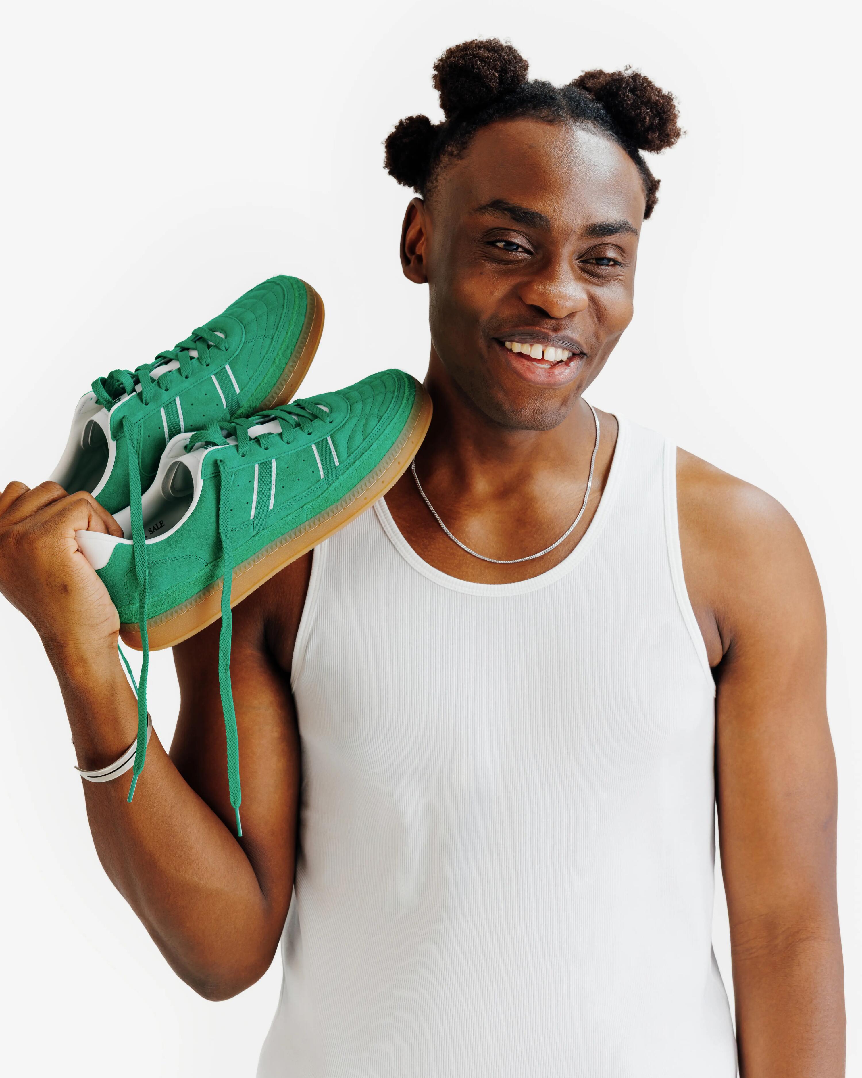 Das Bild zeigt einen jungen Mann vor einem schlichten, weißen Hintergrund, der ein Paar leuchtend grüne Sneaker aus Wildleder präsentiert. Er trägt ein weißes, geripptes Tanktop und hält die Schuhe lässig an den Schnürsenkeln über seine Schulter. Sein Blick ist zur Seite gesenkt, was dem Bild eine moderne und modische Ästhetik verleiht. Die braune Gummisohle und die weißen Akzente der Sneaker kontrastieren stark mit dem kräftigen Grün des Obermaterials.