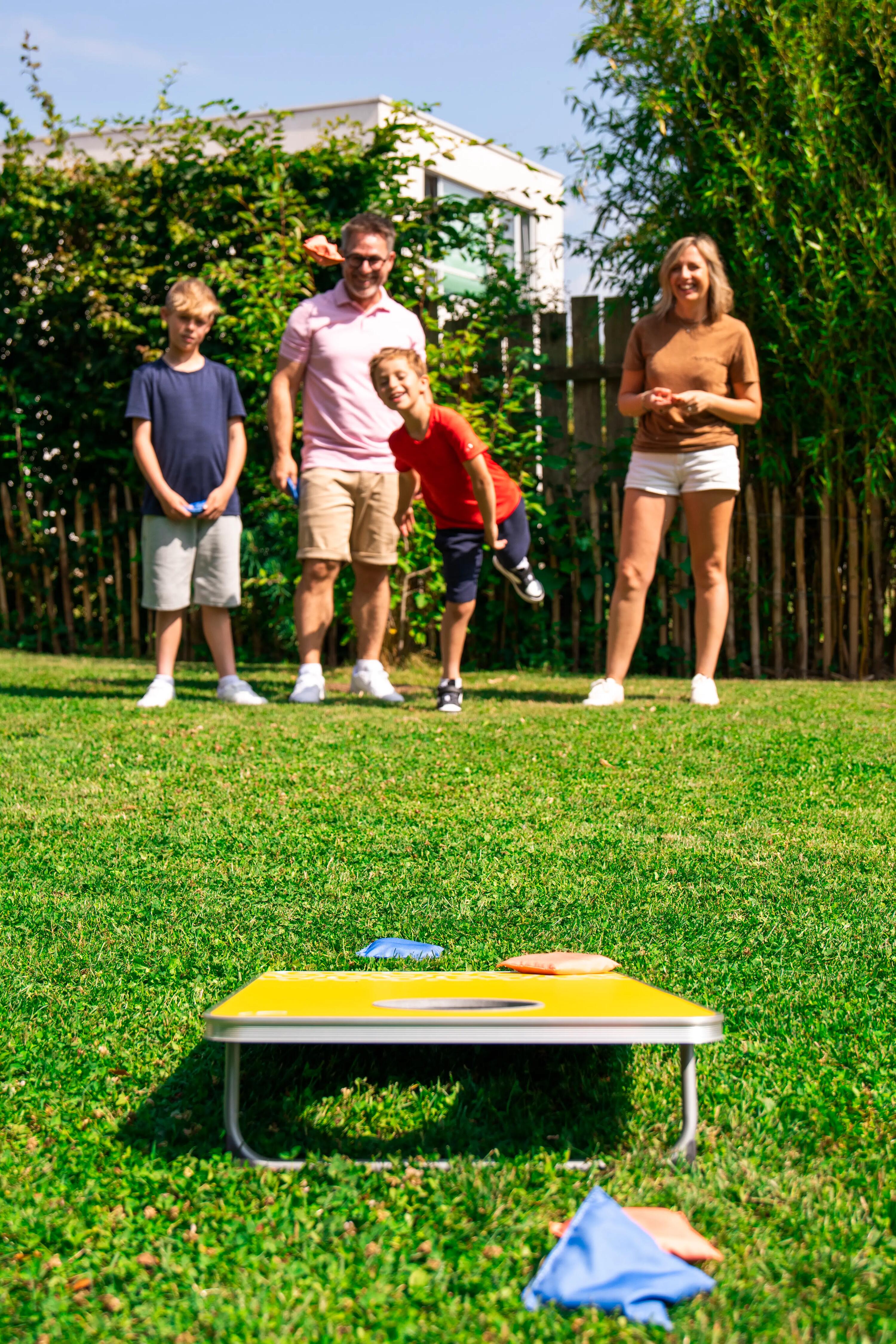 Eine Familie spielt gemeinsam Cornhole im sonnigen Garten, während ein Junge im roten Shirt einen Säckchen auf das gelbe Board wirft.