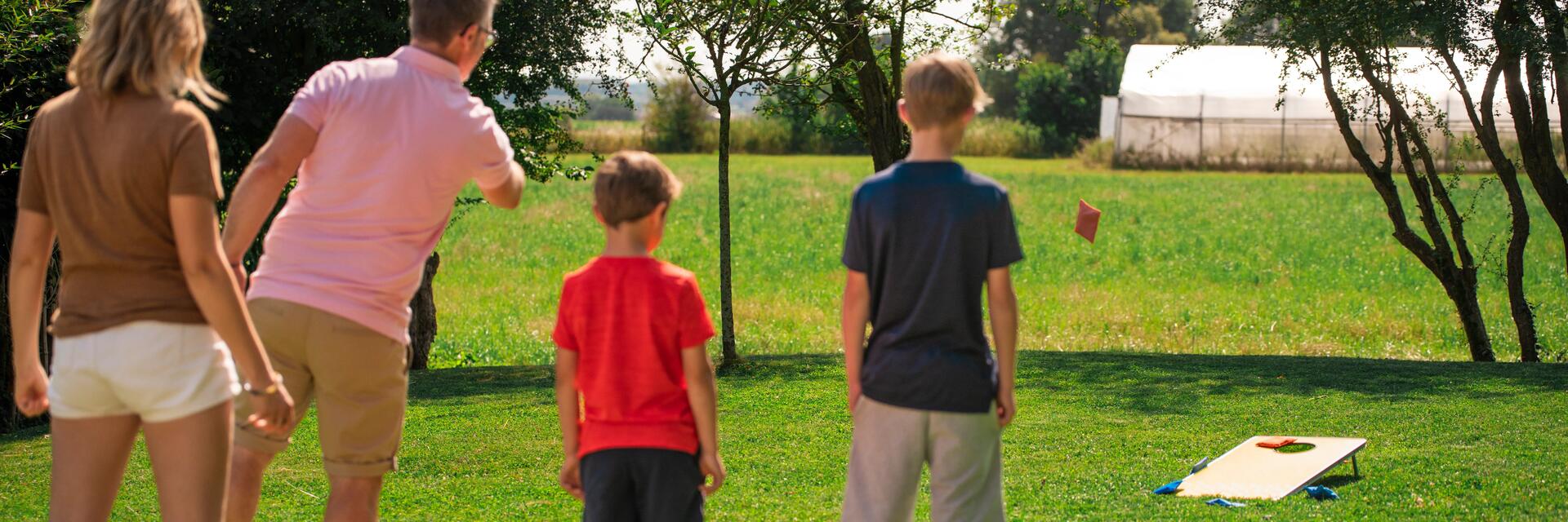 Eine Familie steht auf einer grünen Wiese und spielt Cornhole, wobei ein Mann im rosa Poloshirt gerade ein Säckchen in Richtung des hölzernen Zielbretts wirft.