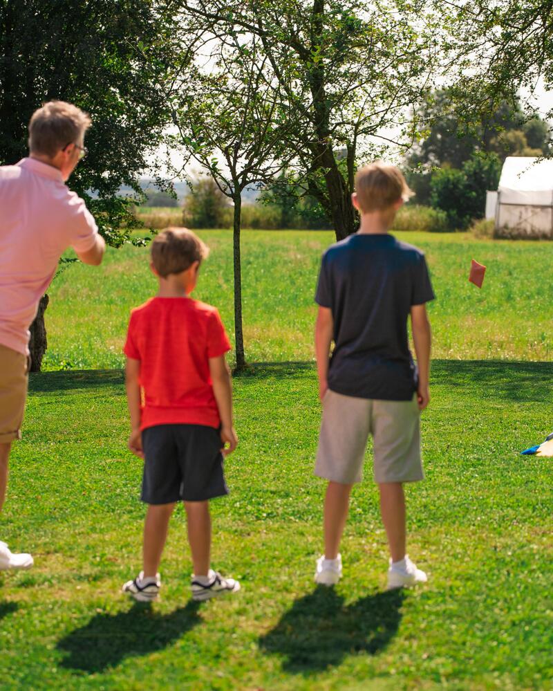 Eine Familie steht auf einer grünen Wiese und spielt Cornhole, wobei ein Mann im rosa Poloshirt gerade ein Säckchen in Richtung des hölzernen Zielbretts wirft.