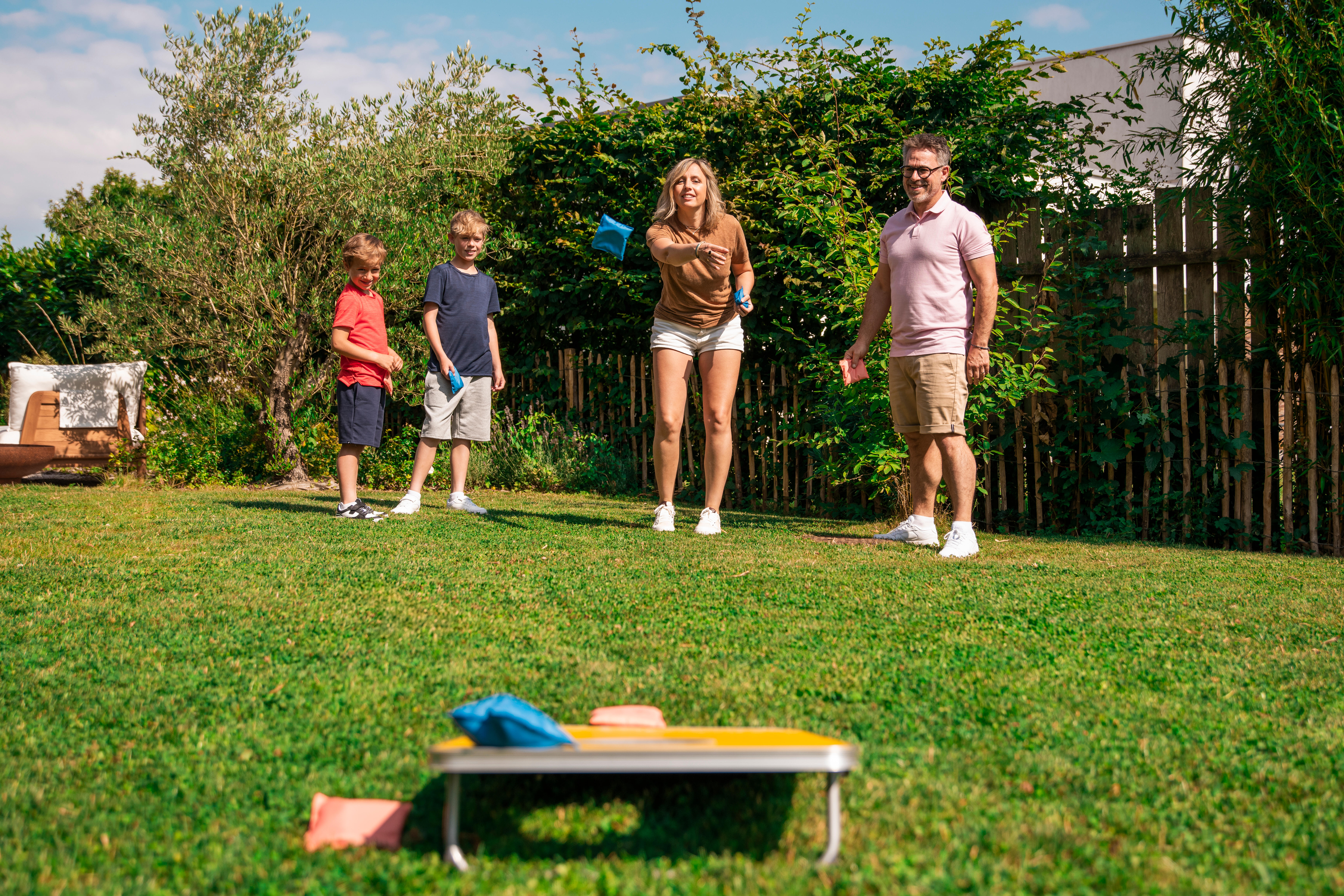 Family throwing bean bags toward yellow cornhole board in garden
