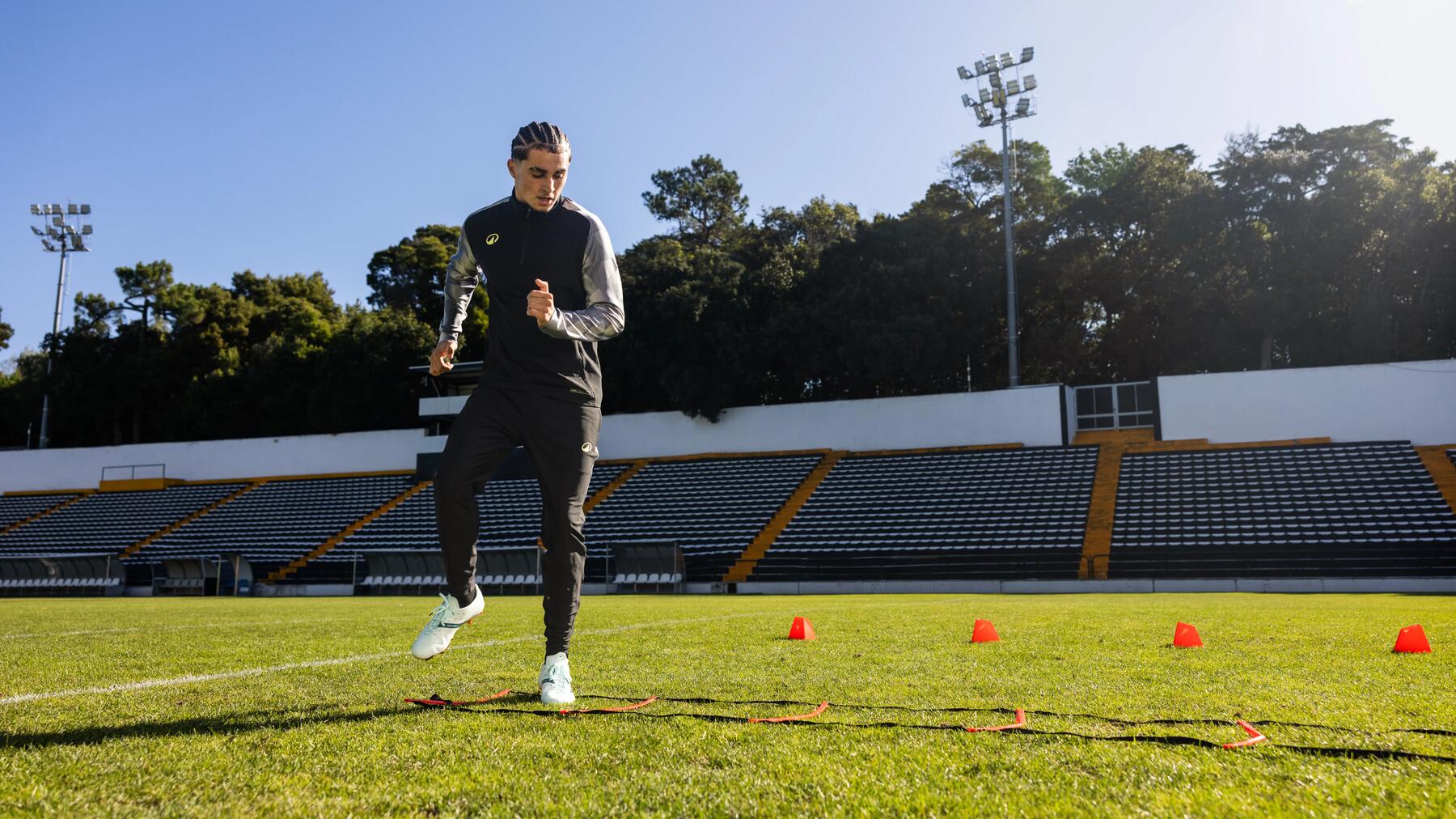 Ein junger Mann absolviert auf einem Rasenplatz vor leeren Tribünen ein Lauf- und Koordinationsleiter-Training bei Sonnenschein.