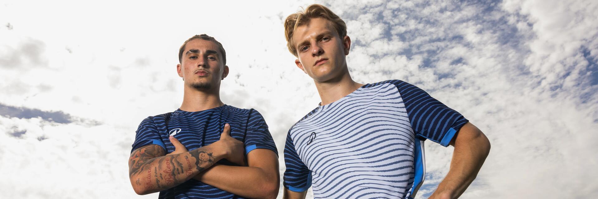 Zwei junge Männer in blau-weiß geschmusterten Sportshirts stehen im Freien vor leicht bewölktem Himmel und blicken selbstbewusst in die Kamera.