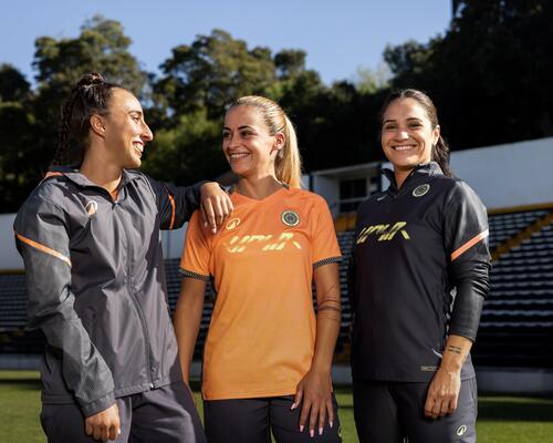 Drei lächelnde Fußballerinnen in Sportkleidung stehen eng zusammen auf einem Spielfeld vor den Zuschauerrängen eines Stadions.