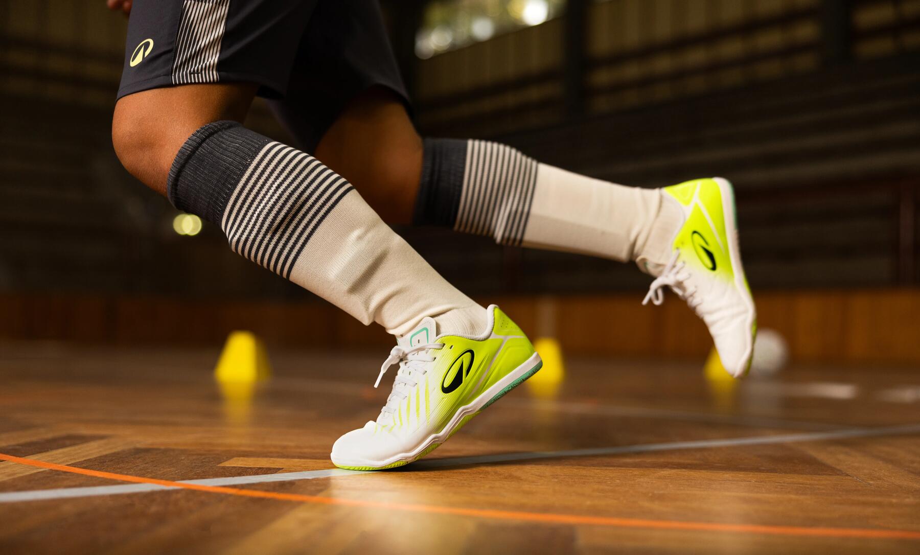 Nahaufnahme der Beine eines Futsal-Spielers in einer Sporthalle, der weiß-gelbe Hallenschuhe und gestreifte Stutzen trägt.
