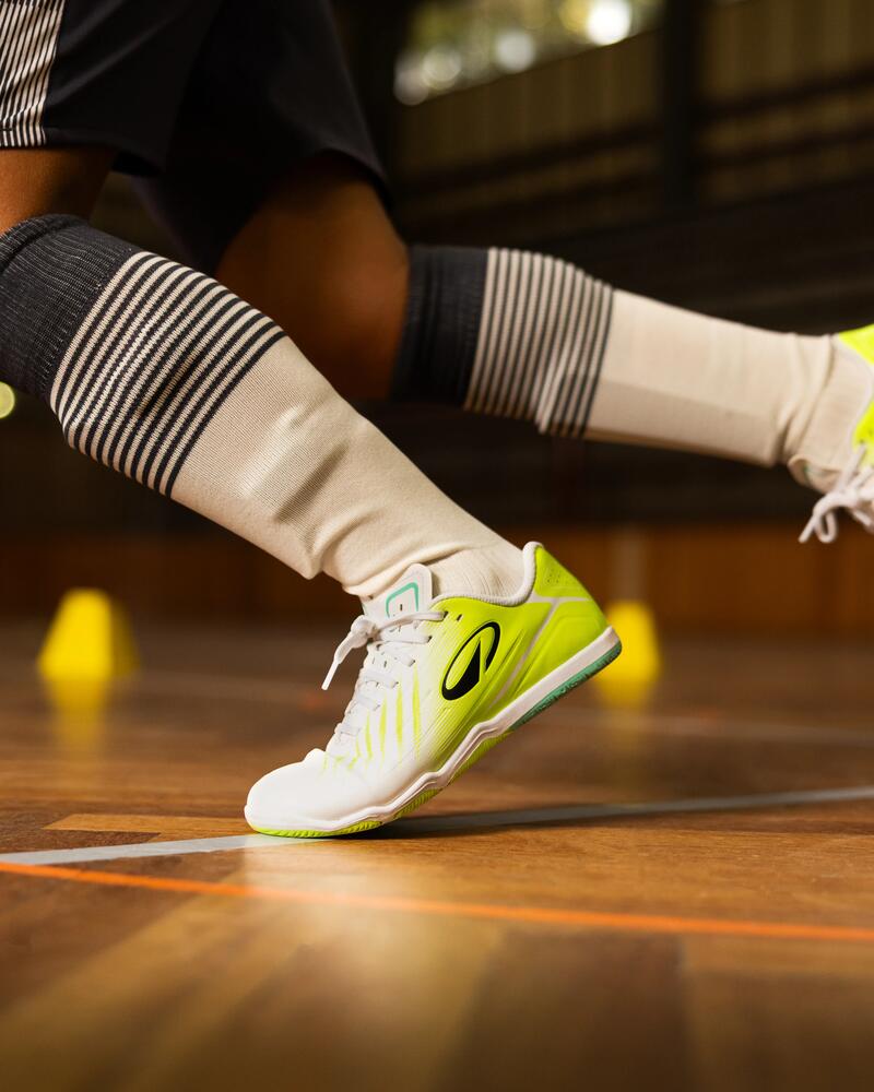 Eine Nahaufnahme aus bodennaher Perspektive zeigt die dynamisch laufenden Beine einer Person in einer Sporthalle, die gestreifte Kniestrümpfe und auffällige weiß-neongelbe Hallensportschuhe trägt, während im Hintergrund kleine gelbe Trainingshütchen auf dem Holzparkett stehen.