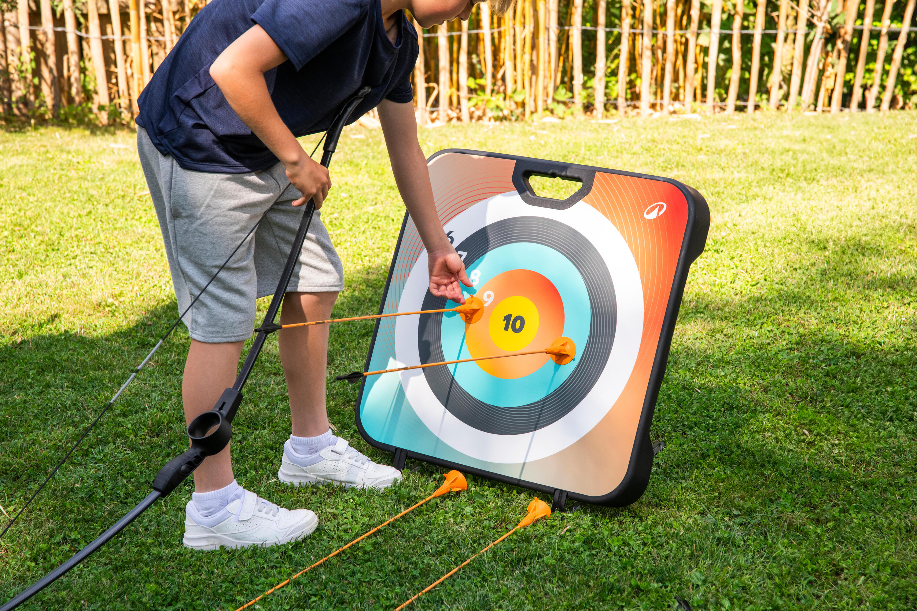 Alt-Text: Nahaufnahme eines Jungen in kurzen Hosen und T-Shirt, der auf einem Rasen steht und einen Pfeil aus einer Spielzeug-Bogenschießscheibe zieht. Die Scheibe ist farbig markiert mit Zahlen, und zwei Pfeile stecken bereits darin.