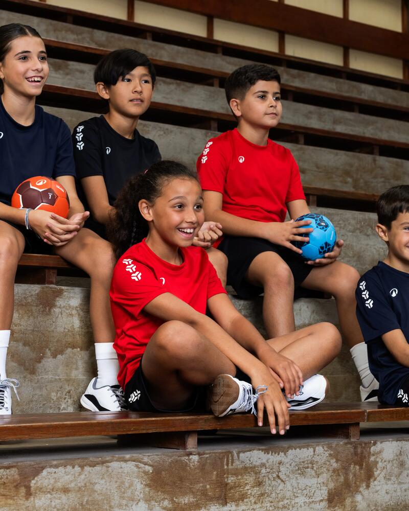 Fünf Kinder sitzen in verschiedenenfarbigen Trikots auf einer Tribüne und tragen dabei Kipsta Handball Klamotten. Zwei von ihnen haben einen Handball in der Hand.