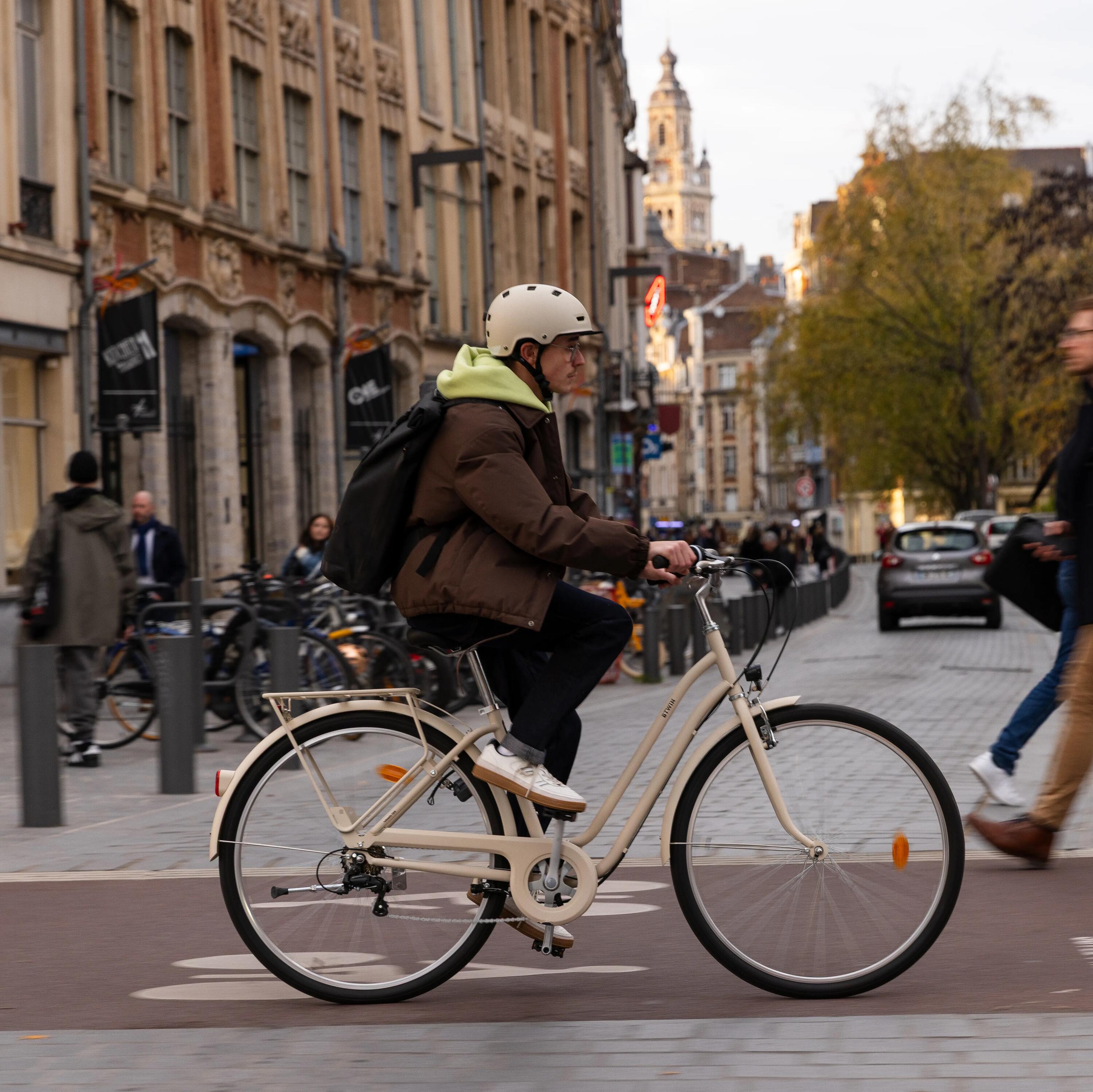 Ein junger Mann mit hellem Helm, brauner Jacke und Rucksack fährt ein cremefarbenes Hollandrad der Marke Elops auf einem Radweg in einer historischen europäischen Stadtstraße. Der Radfahrer im Vordergrund ist scharf, während der Hintergrund (Gebäude mit 'Monoprix'-Schild, Passanten, ein Auto und Straßenschilder) durch Bewegungsunschärfe Dynamik und Geschwindigkeit vermittelt. Die Szene ist bei Tageslicht und fängt das urbane Pendeln ein.
