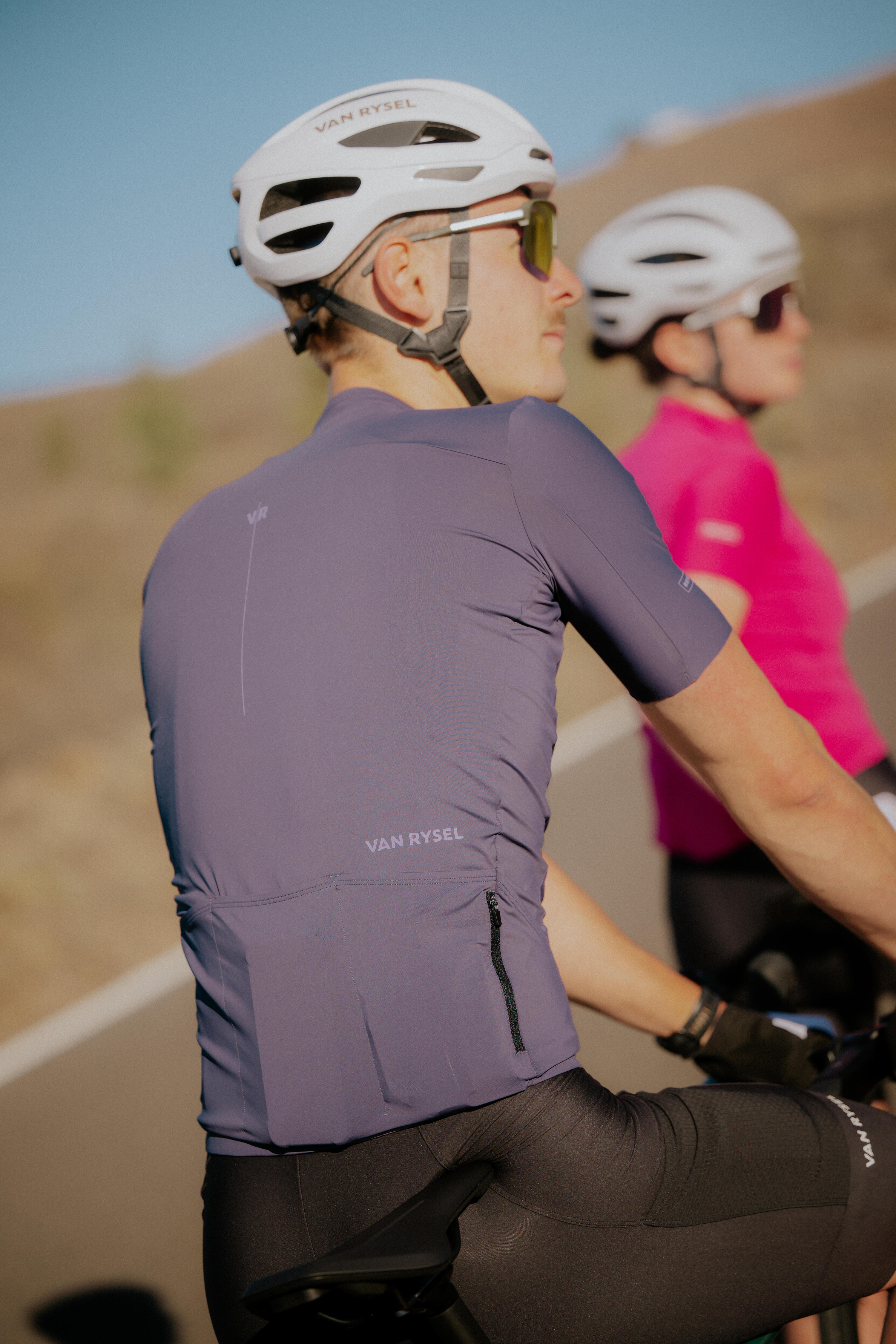 Zwei Rennradfahrer – ein Mann im Vordergrund in lila-grauem Outfit und eine Frau im Hintergrund in Pink – fahren bei Sonnenschein auf einer Landstraße. Beide tragen weiße Van Rysel Helme und schauen aufmerksam über die Schulter nach vorne.