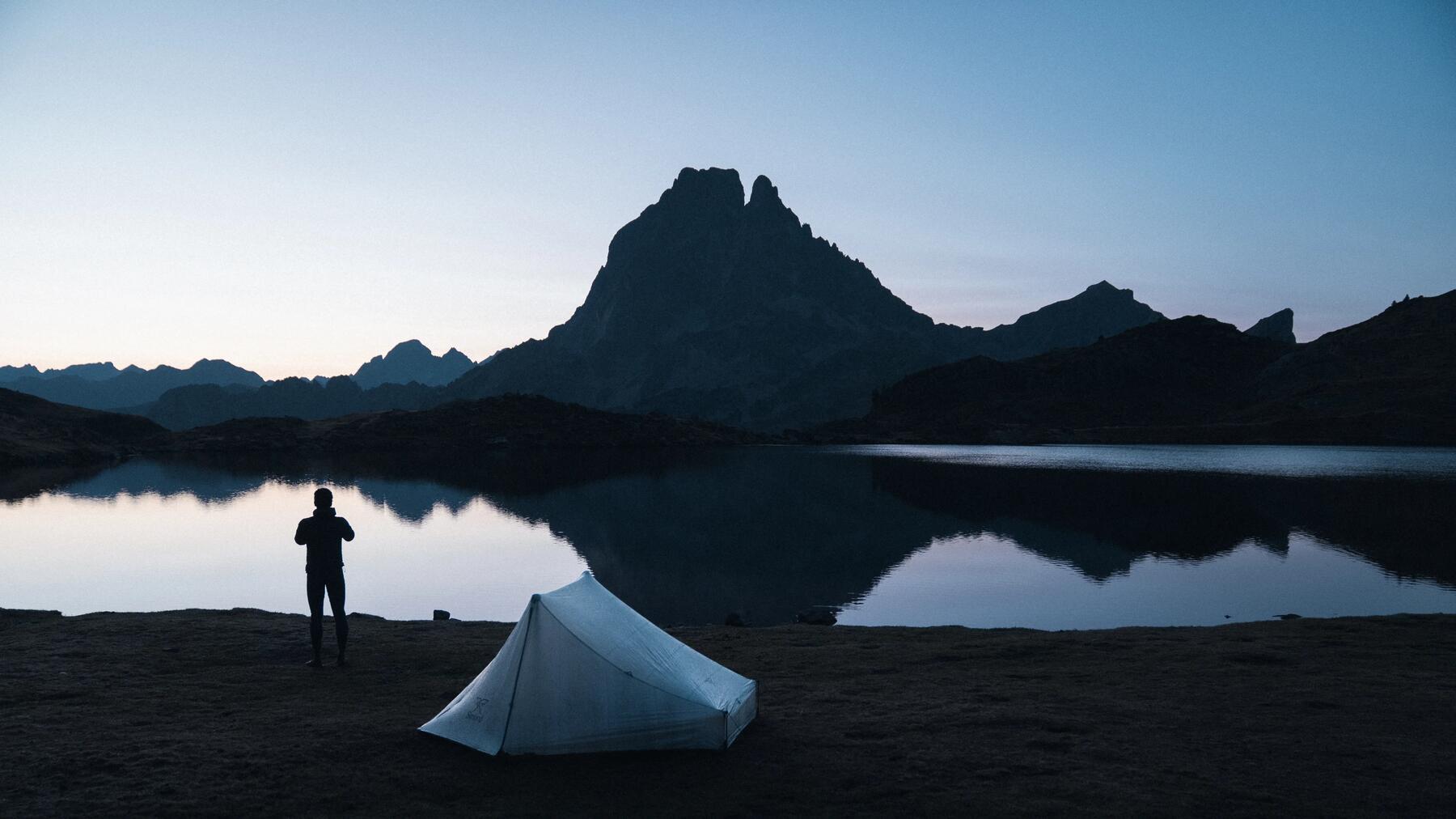 Ein Mann steht im Dunkeln vor seinem Zelt am Bergsee und muss es flicken