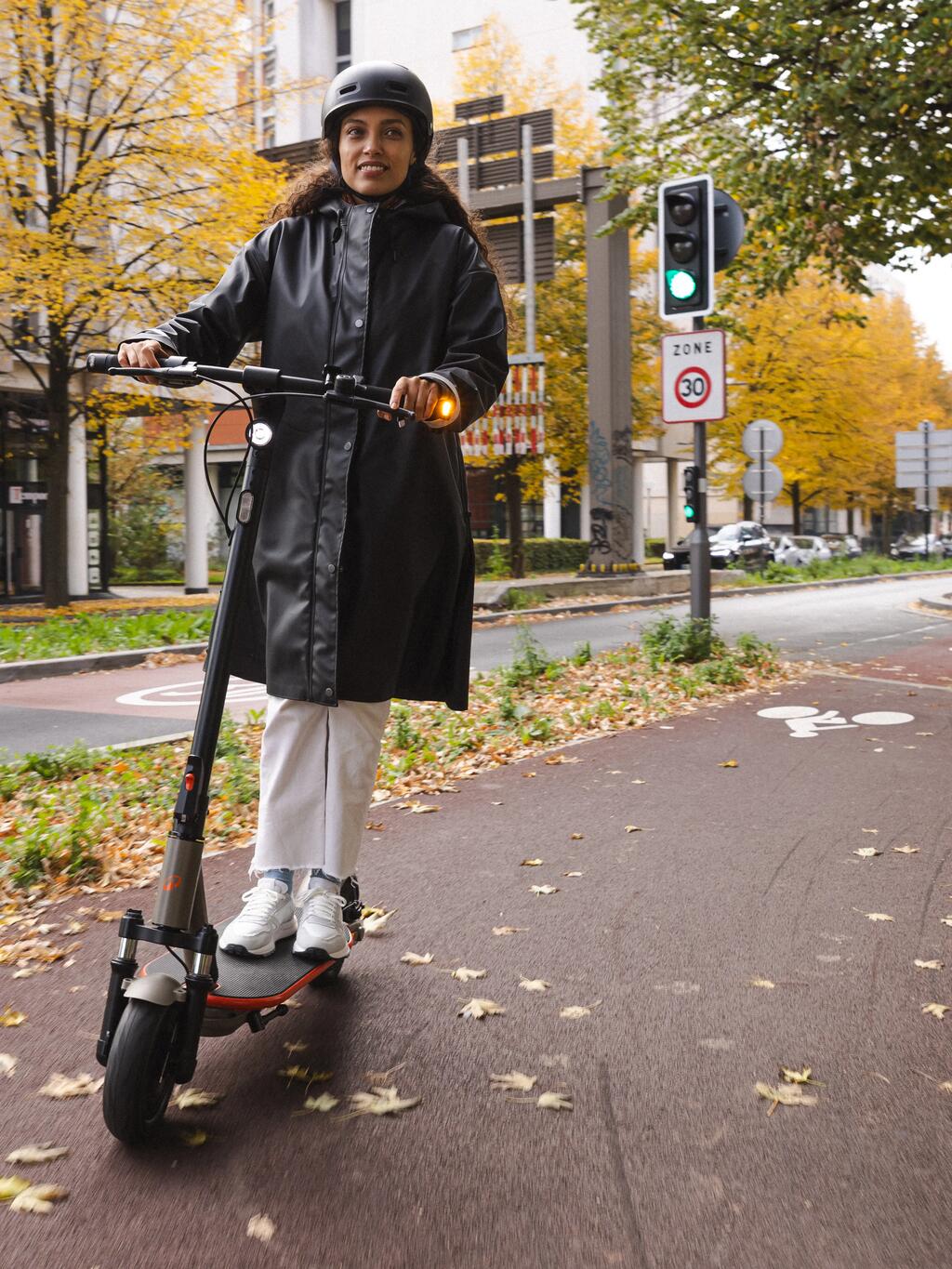 Eine Frau mit Helm fährt auf einem elektrischen Tretroller (E-Scooter) auf einem Gehweg.