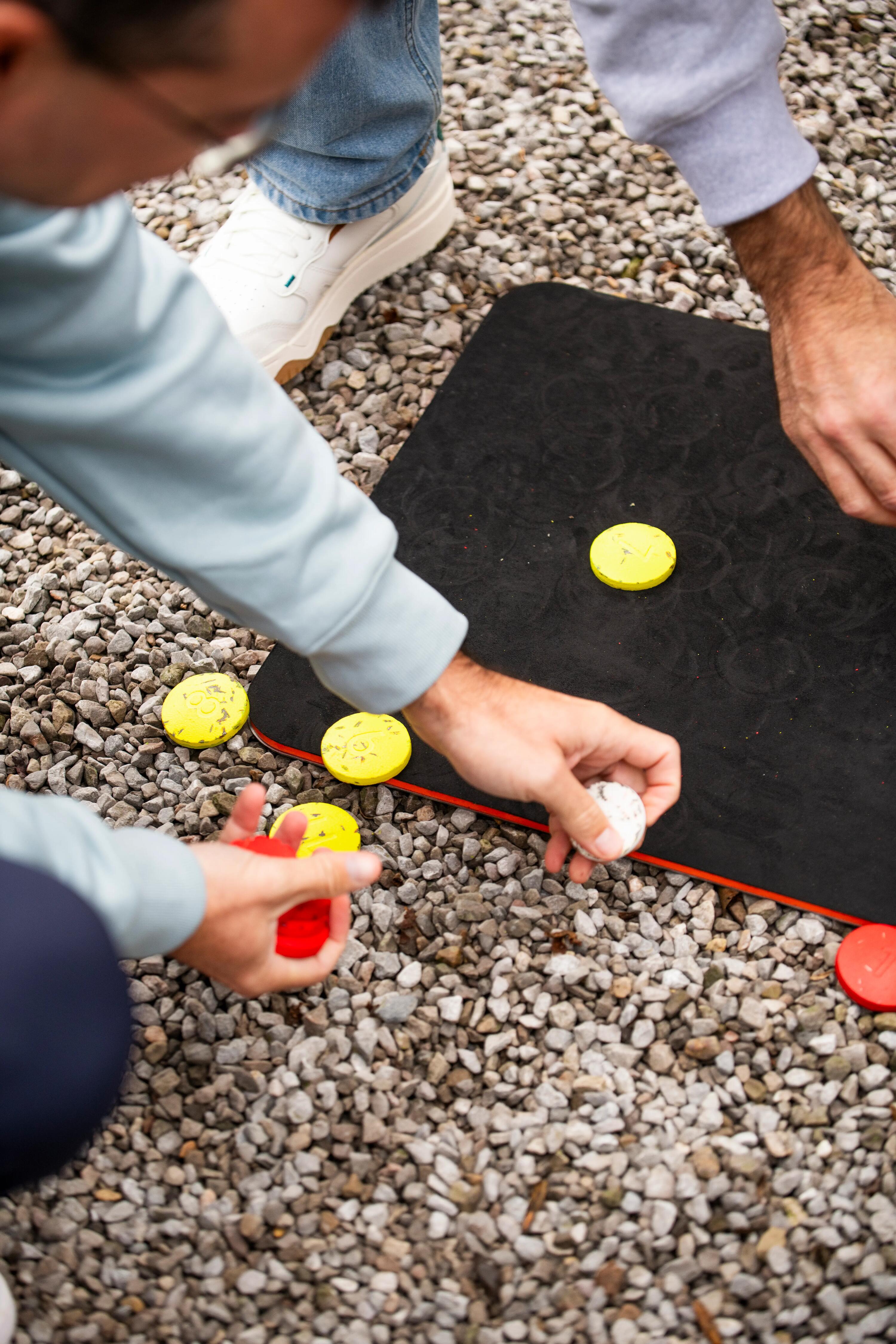 Zwei Personen spielen Palet auf einem Kiesuntergrund mit einer schwarzen Spielmatte und bunten Holzscheiben.