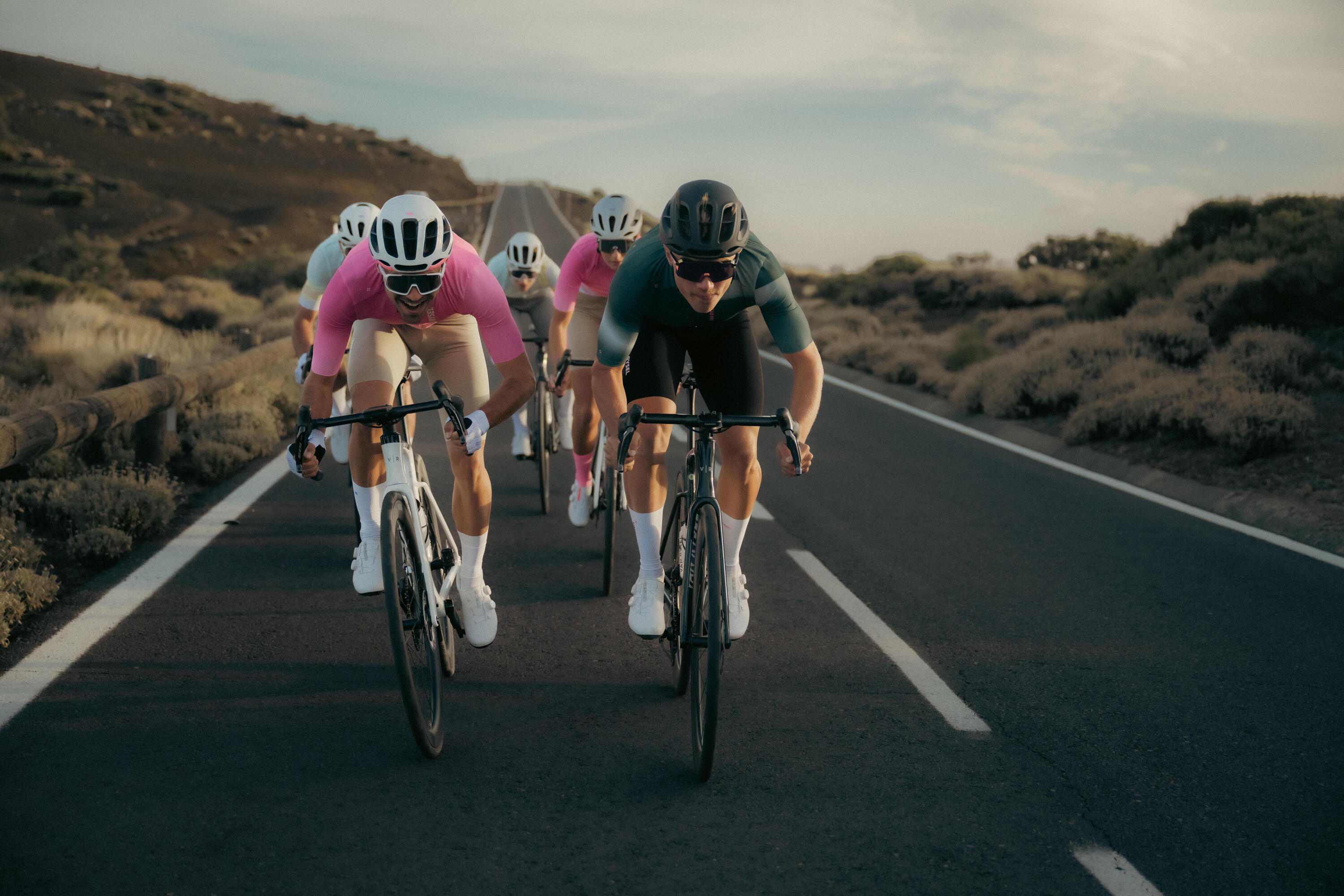 Eine Gruppe von Rennradfahrern in aerodynamischer Position fährt auf einer Asphaltstraße durch eine karge Hügellandschaft. Im Fokus stehen zwei Fahrer im Vordergrund – einer in einem pinkfarbenen und einer in einem dunkelgrünen Trikot.