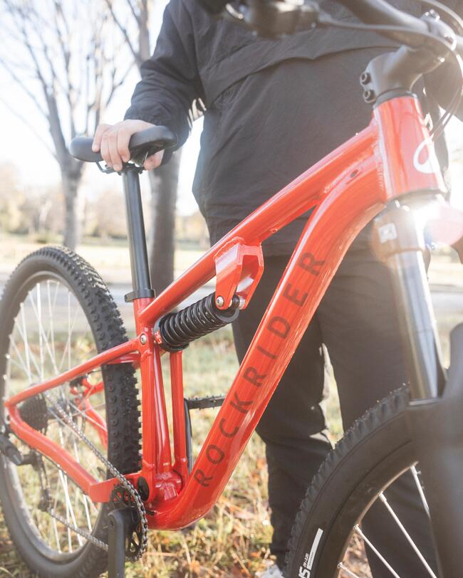 Ein Foto eines roten, vollgefederten Rockrider Mountainbikes, das im Freien auf Gras steht, mit einem unscharfen Hintergrund aus blattlosen Bäumen und warmem Sonnenlicht. Ein Mann in einer schwarzen Jacke steht hinter dem Fahrrad und hält es am Sattel fest.