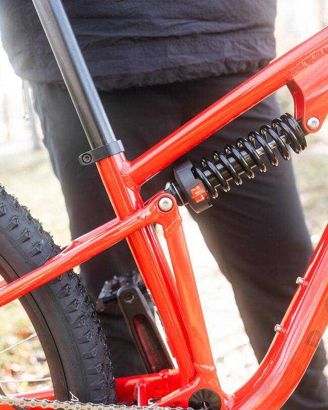 Nahaufnahme des roten Rahmens eines vollgefederten Mountainbikes mit Fokus auf das schwarze Stahlfederbein und den Antriebsbereich. Im Hintergrund steht eine Person in dunkler Kleidung und hellen Sneakern auf einem herbstlichen Waldweg.