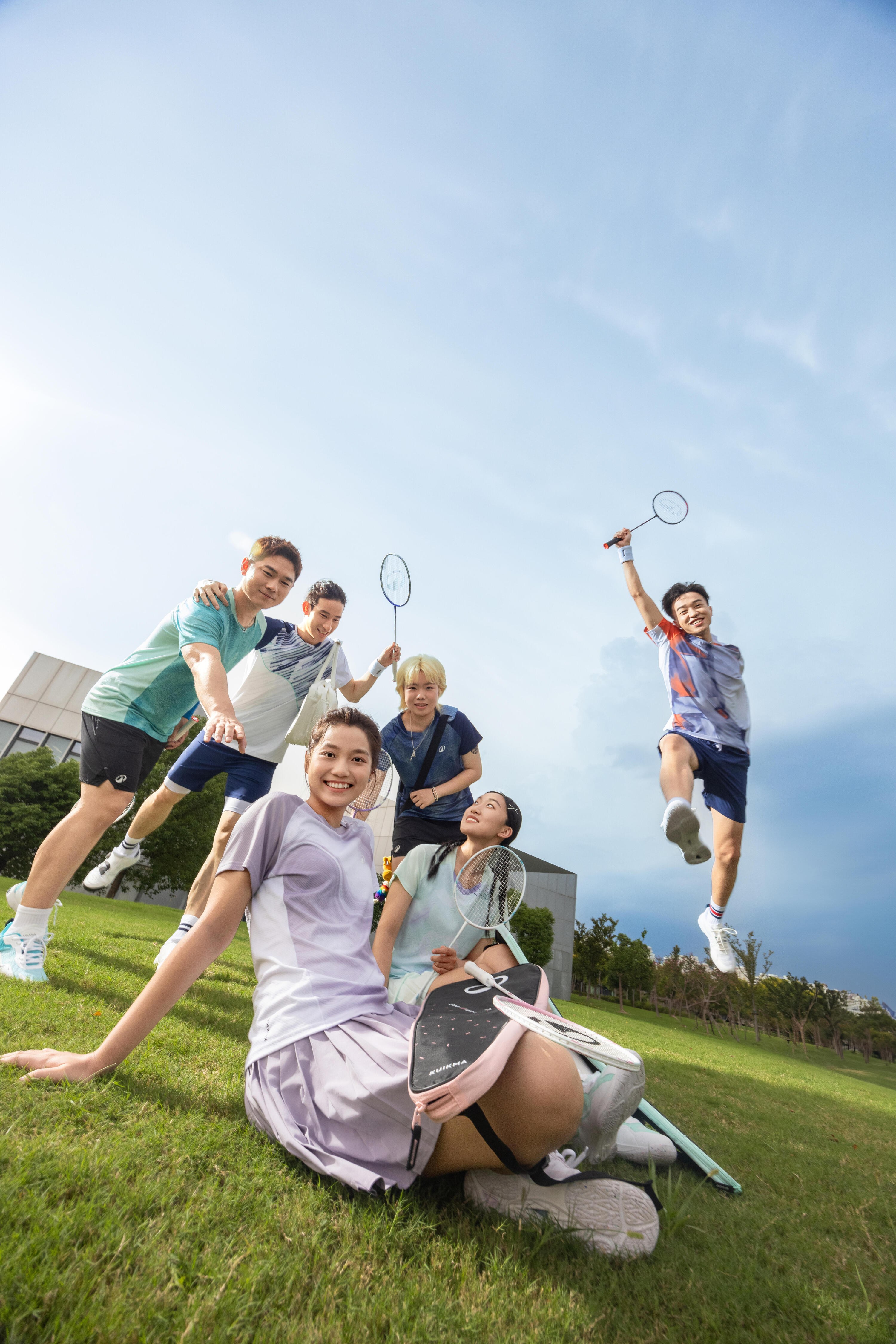 Eine Gruppe junger, lachender Badmintonspieler posiert ausgelassen auf einer grünen Wiese; eine Spielerin im Vordergrund sitzt entspannt im Gras, während ein Mitspieler im Hintergrund mit erhobenem Schläger in die Luft springt.