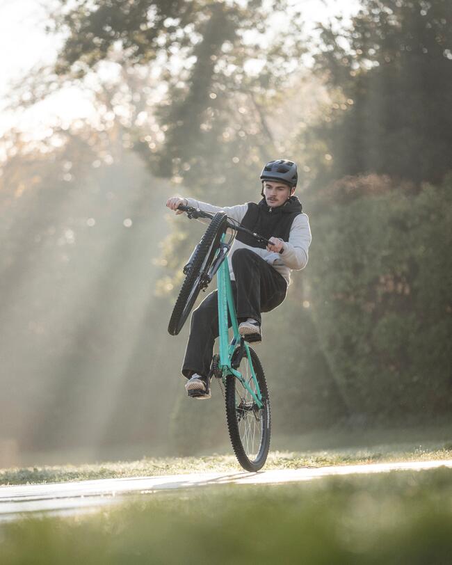 Ein Mann mit Helm vollführt auf einem türkisfarbenen Mountainbike einen Wheelie auf einem Asphaltweg. Die Szenerie ist in warmes Morgenlicht mit sichtbaren Sonnenstrahlen durch die Bäume getaucht.