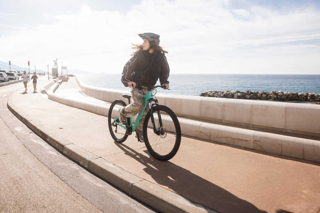 Mlada ženska s čelado vozi turkizno gorsko kolo Rockrider po obalni poti in gleda proti morju.