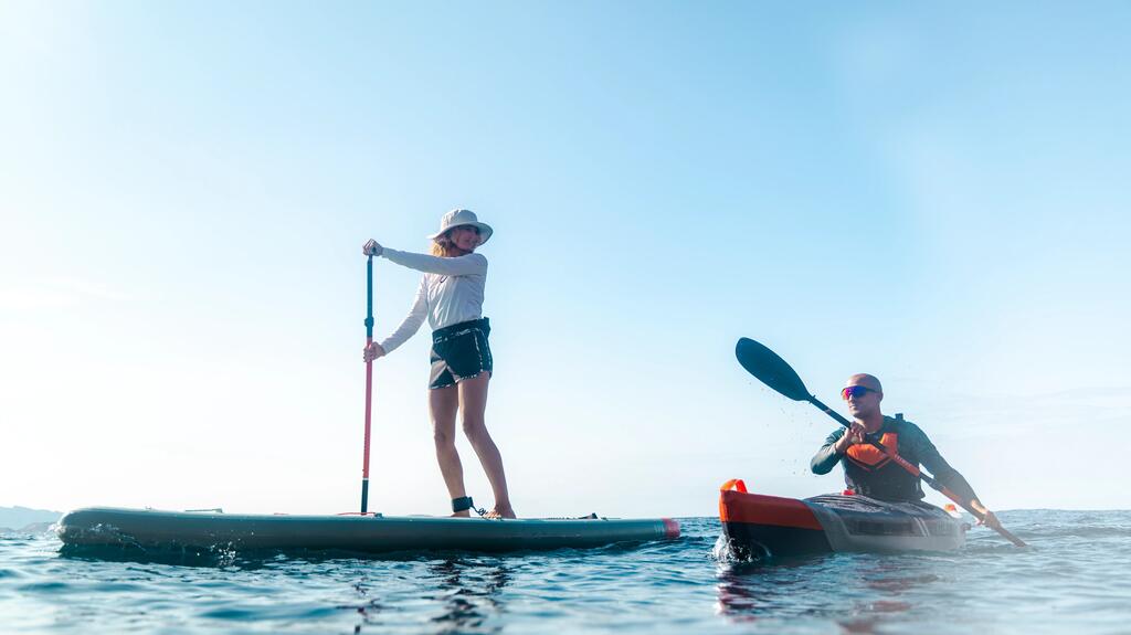Eine Frau steht auf einem Stand-Up-Paddleboard und hält ein Paddel, während rechts neben ihr ein Mann in einem Kajak durch das ruhige, blaue Meer paddelt. Beide tragen Sportbekleidung und Kopfbedeckungen bzw. eine Sonnenbrille unter einem klaren, hellen Himmel.