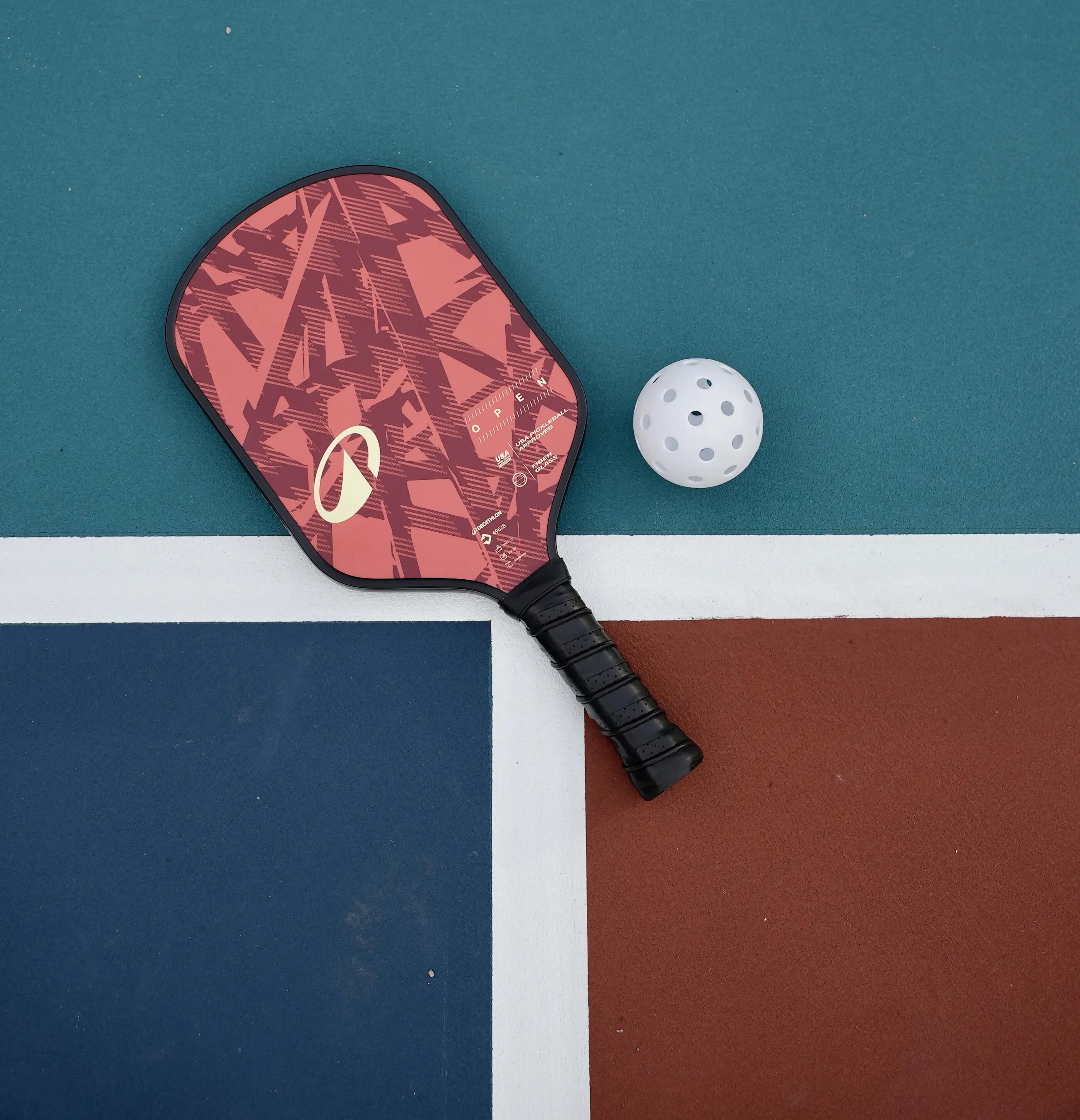 Ein rot gemusterter Pickleball-Schläger und ein weißer Lochball liegen auf dem farbigen Boden eines Spielfelds.