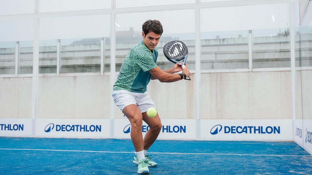 Ein Mann auf einem blauen Padel-Court holt mit einem Padel-Schläger zweihändig zum Schlag auf einen gelben Ball aus. Im Hintergrund sind Decathlon-Sponsorings an den Glaswänden sichtbar.
