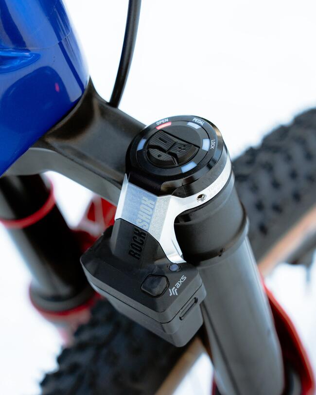 Grande plano de uma bicicleta de montanha Rockrider azul e preta com letras em verde, uma suspensão RockShox vermelha e pneus com paredes laterais castanhas, sobre fundo branco.