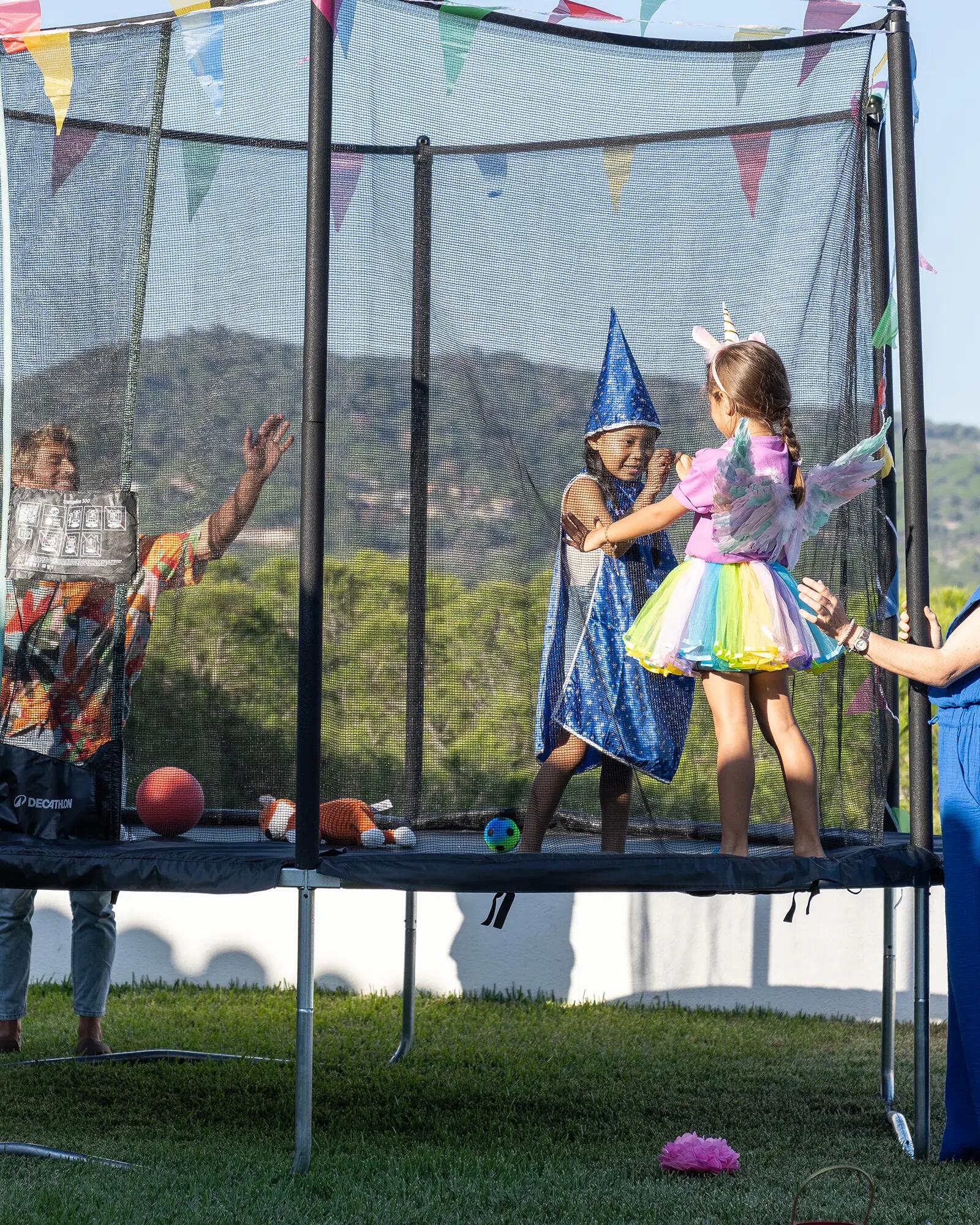 Zwei Kinder in Kostümen spielen auf einem Trampolin im Freien, beobachtet von zwei Erwachsenen.