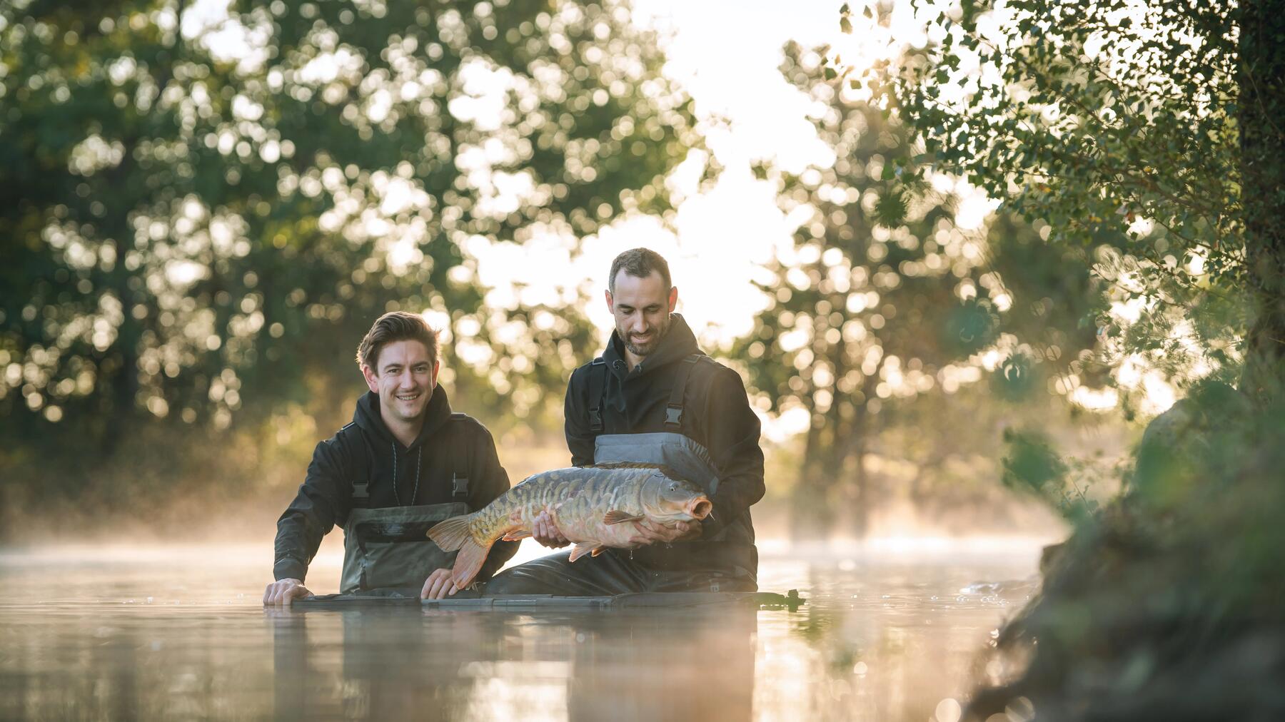 Zwei Angler stehen mit Wathose im Wasser und einer hält einen gefangenen Karpfen in den Händen