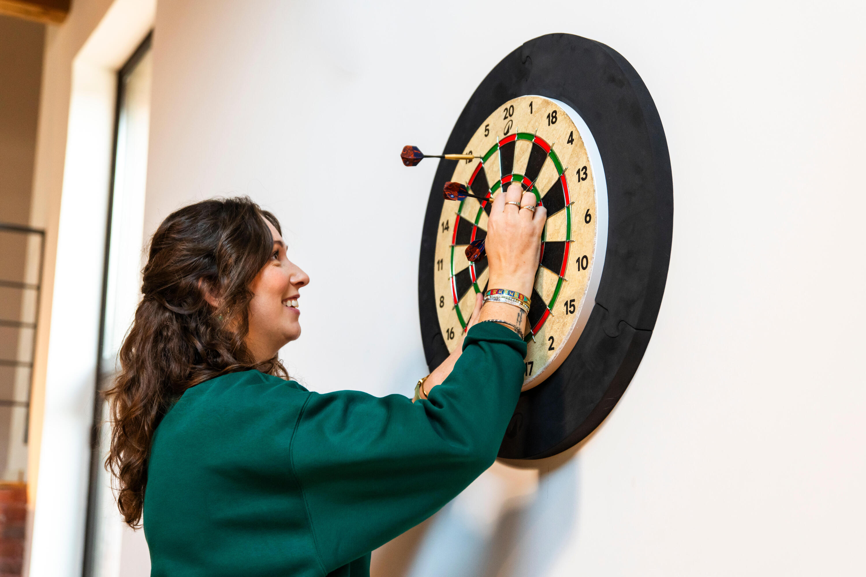 Eine Frau zieht lächend die Dartpfeile aus einer Steeldartscheibe, die an der Wand hängt.