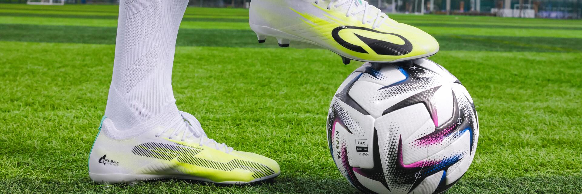 Nahaufnahme der Beine eines Fußballspielers in weißen Stutzen und gelb-weißen Kickschuhen, der auf einem Rasenplatz einen Fuß auf dem Fußball hat.