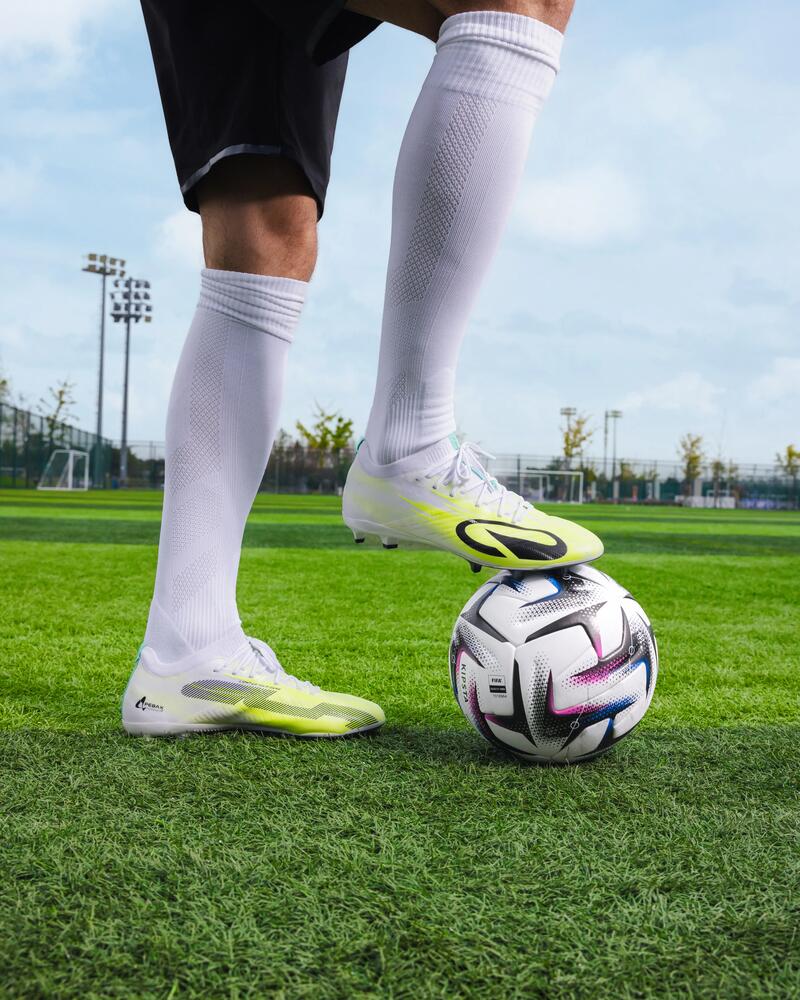 Ein Fußballspieler in weißen Stutzen und gelb-weißen Fußballschuhen steht auf einem grünen Kunstrasenplatz und hat einen Fuß auf einem Fußball platziert.
