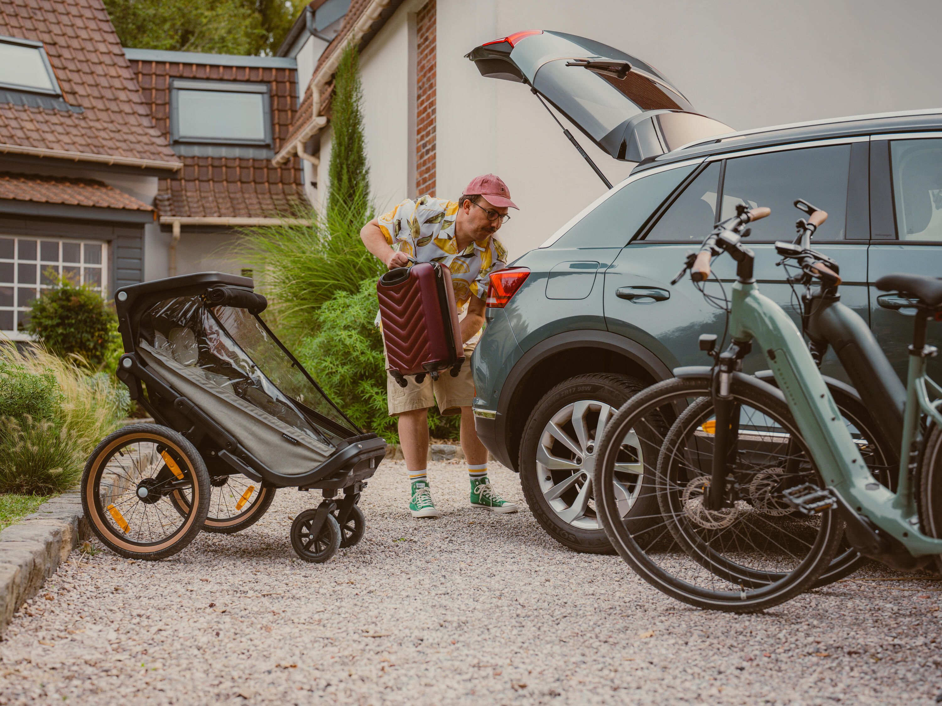 Ein Mann belädt ein Auto für die Urlaubsfahrt. Links im Bild ist der XPL 900 Fahrradanhänger zu sehen, rechts vor dem Auto stehen zwei E-ACTV Fahrräder.