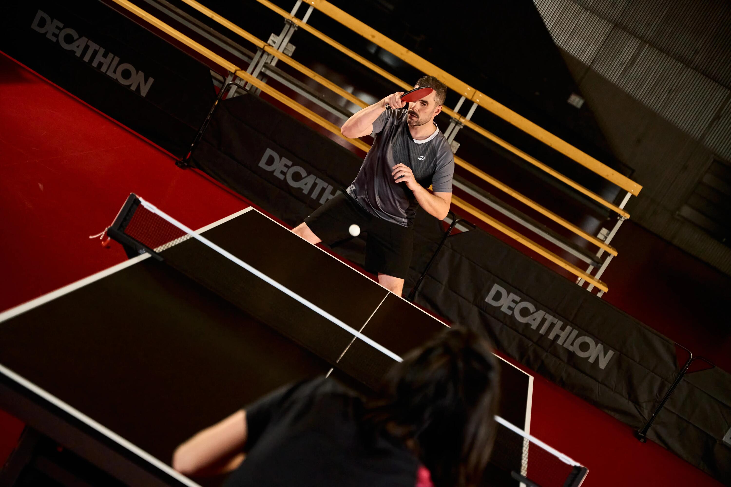Eine dynamische Actionaufnahme aus der Vogelperspektive, aufgenommen während eines Tischtennisspiels in der Halle. Im unmittelbaren Vordergrund beobachtet ein Spieler von hinten den Ballwechsel. Auf der anderen Seite des Netzes hält ein konzentrierter Gegner in grauem Hemd und schwarzen Shorts seinen Schläger hoch und bereitet sich darauf vor, den Ball zu schlagen, der in Bewegung ist. Die Spielfeldbegrenzung besteht aus schwarzen Barrieren mit dem sichtbaren 'DECATHLON'-Logo. Der Spielbereich ist durch einen roten Boden und eine dunkle Tischoberfläche definiert, mit gelben Metallstrukturen im Hintergrund.