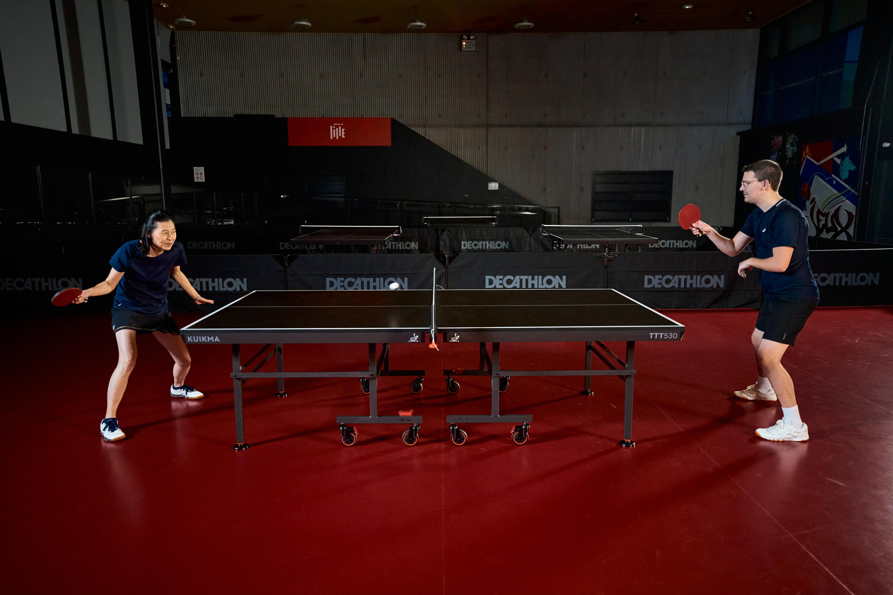Ein Mann und eine Frau spielen engagiert Tischtennis auf einer TTT530-Tischtennisplatte in einer Sporthalle.