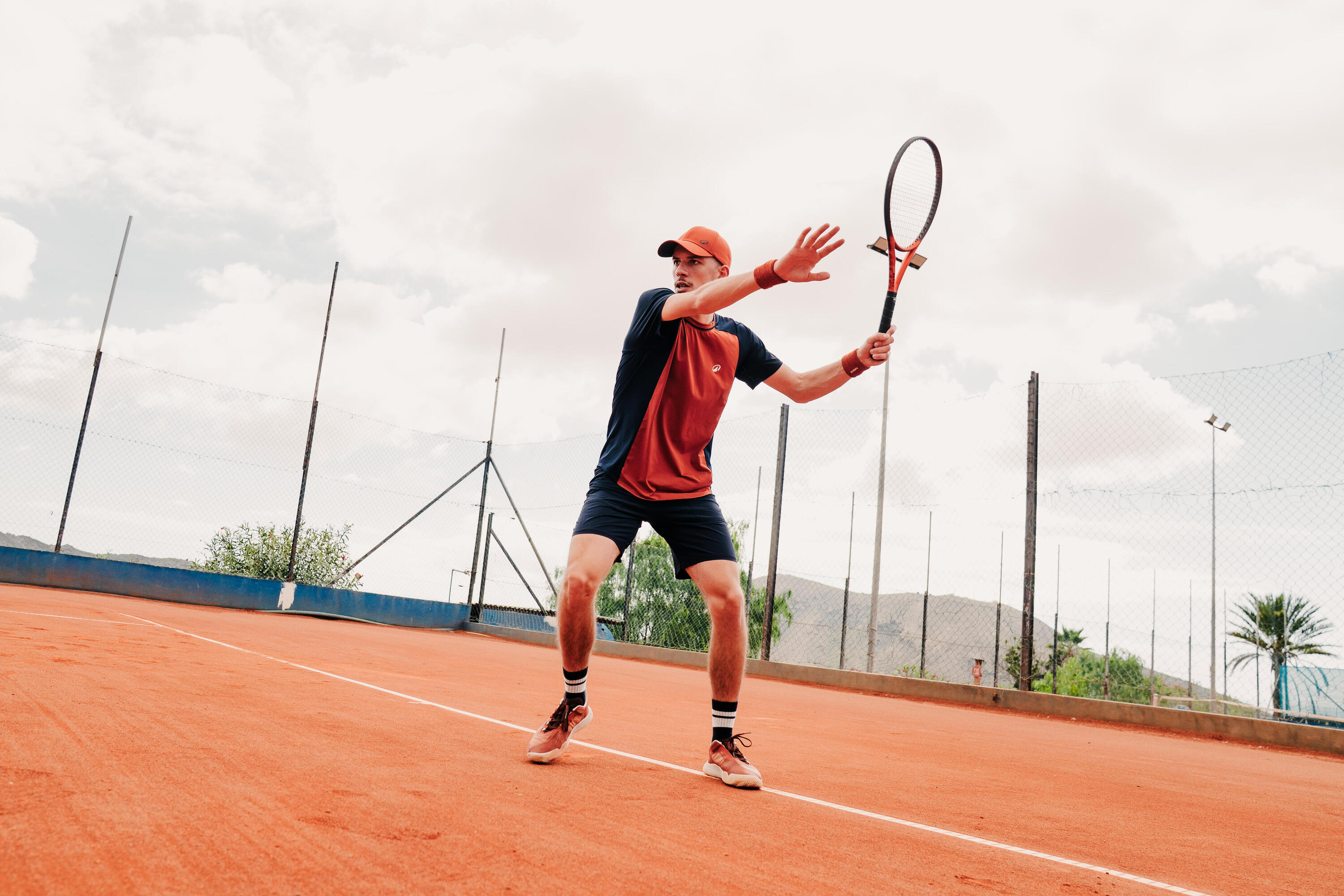 Ein Tennisspieler in konzentrierter Haltung bereitet sich auf einem Sandplatz mit erhobenem Schläger auf einen Schlag vor. Er trägt sportliche Kleidung und eine orangefarbene Kappe vor einer weitläufigen Platzkulisse.