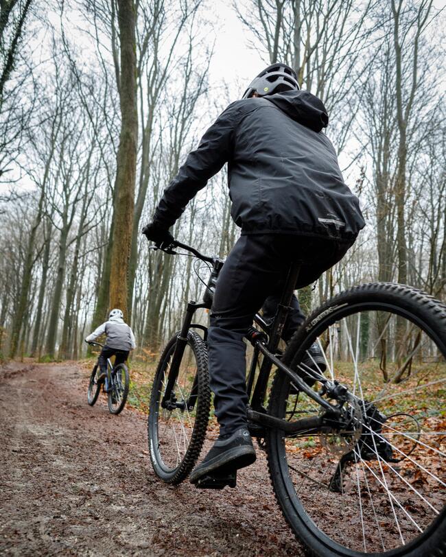 Eine Fotografie aus einer niedrigen Perspektive zeigt einen Mountainbiker in schwarzer Kleidung von hinten, der auf einem matschigen Waldweg fährt. Weiter vorne auf dem von kahlen Bäumen gesäumten Pfad ist ein zweiter Radfahrer zu sehen.